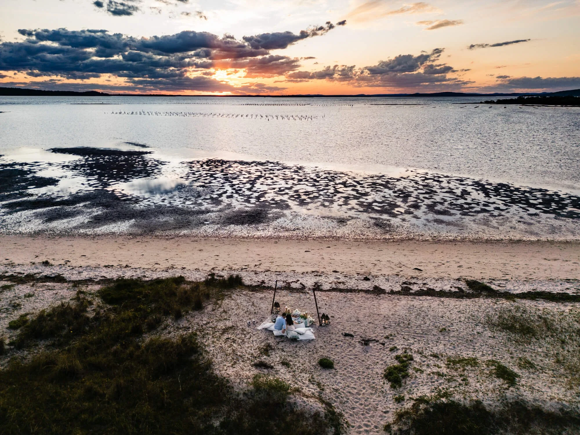 Aerial drone view of a couple at a styled beach picnic on a sandy shoreline at sunset, tidal flats and moody clouds beyond, Soldiers Point, Port Stephens.