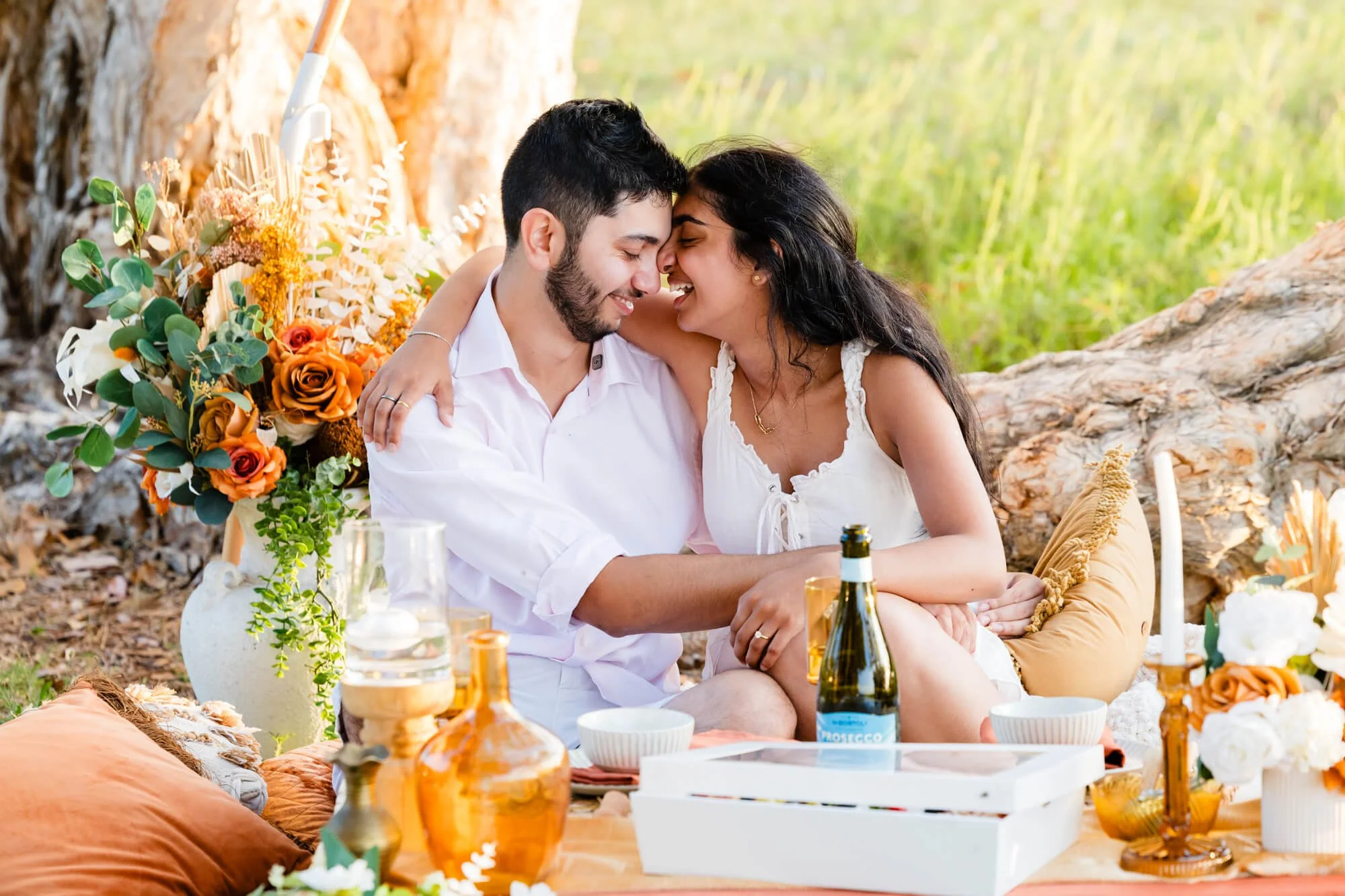 Couple embracing and smiling over a styled picnic with an orange floral arrangement, amber glassware and a bottle of sparkling wine, Soldiers Point, Port Stephens.
