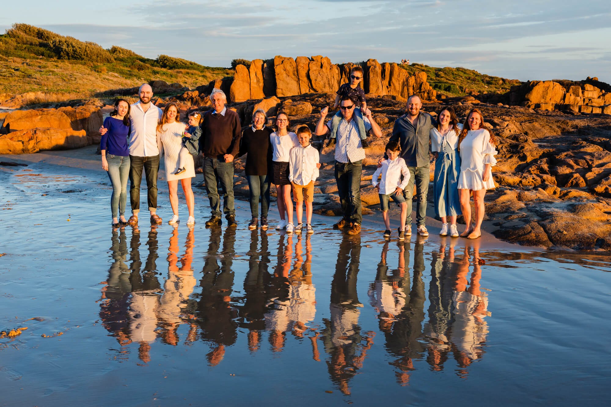 Three-generation family group of about twenty standing together on orange coastal rocks, their reflections in a rock pool at sunset, Birubi Beach, Anna Bay, Port Stephens.