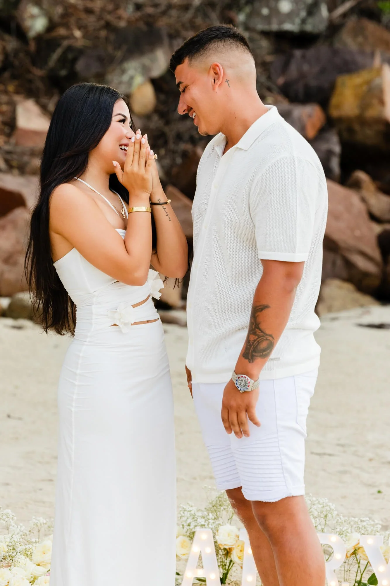 Woman smiling with her hands clasped to her face in happy surprise as her partner looks on, on a rocky beach with flowers set out at their feet, Nelson Bay, Port Stephens.