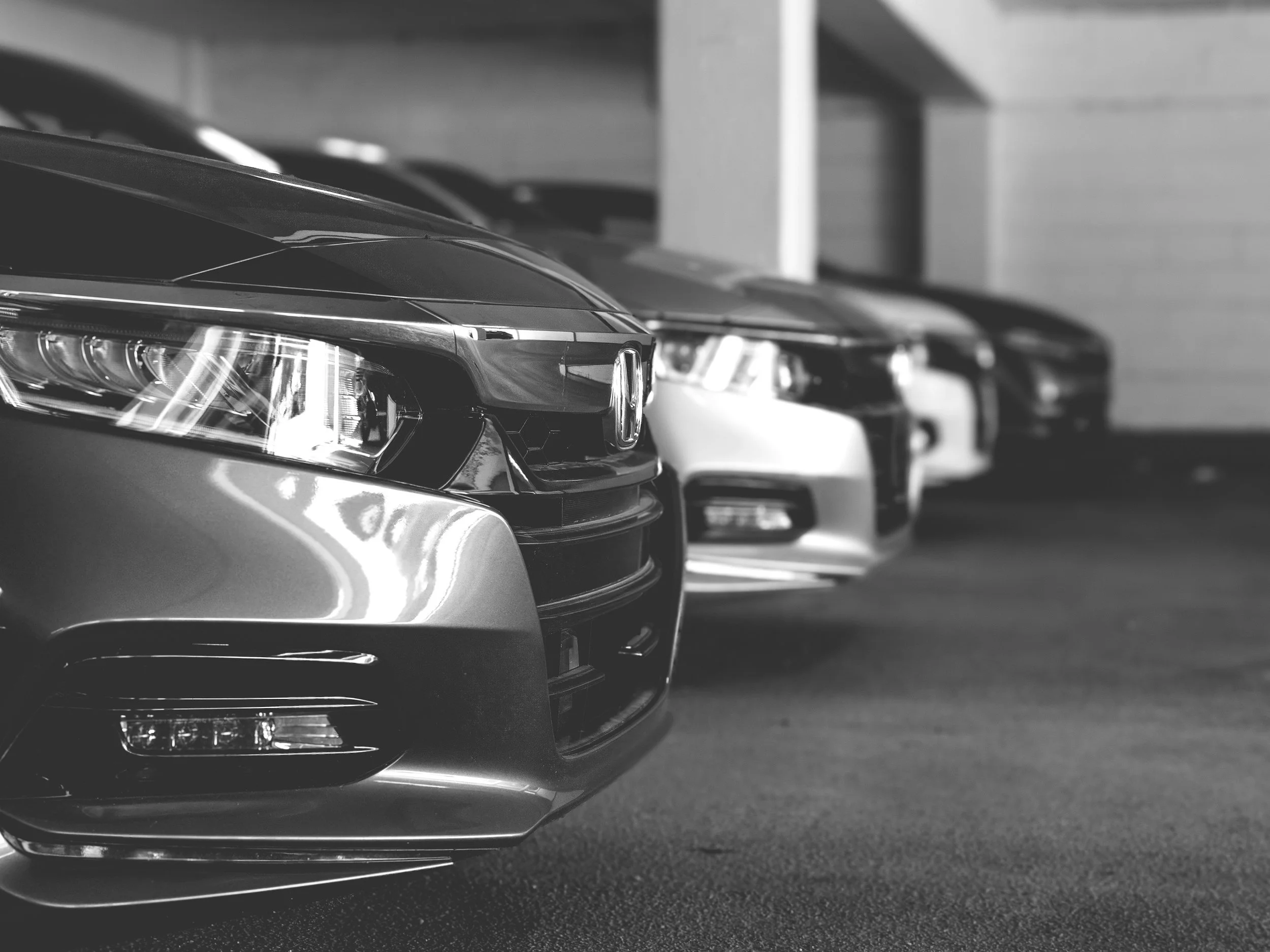 Black and white photograph of several cars parked closely in a parking garage, with a focus on the front part of the vehicles.