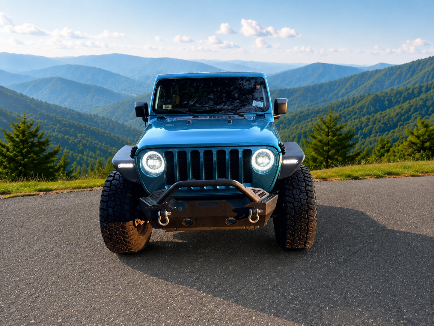 Front view of a blue Jeep parked on a paved road with a mountain range and green trees in the background.