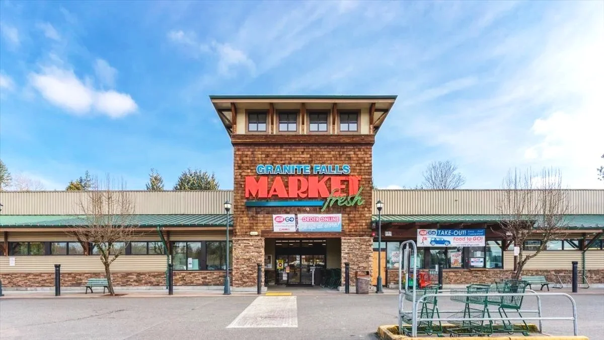 Front view of Granite Falls Market & Fresh grocery store with parking area and shopping carts in the foreground, blue sky with clouds above.