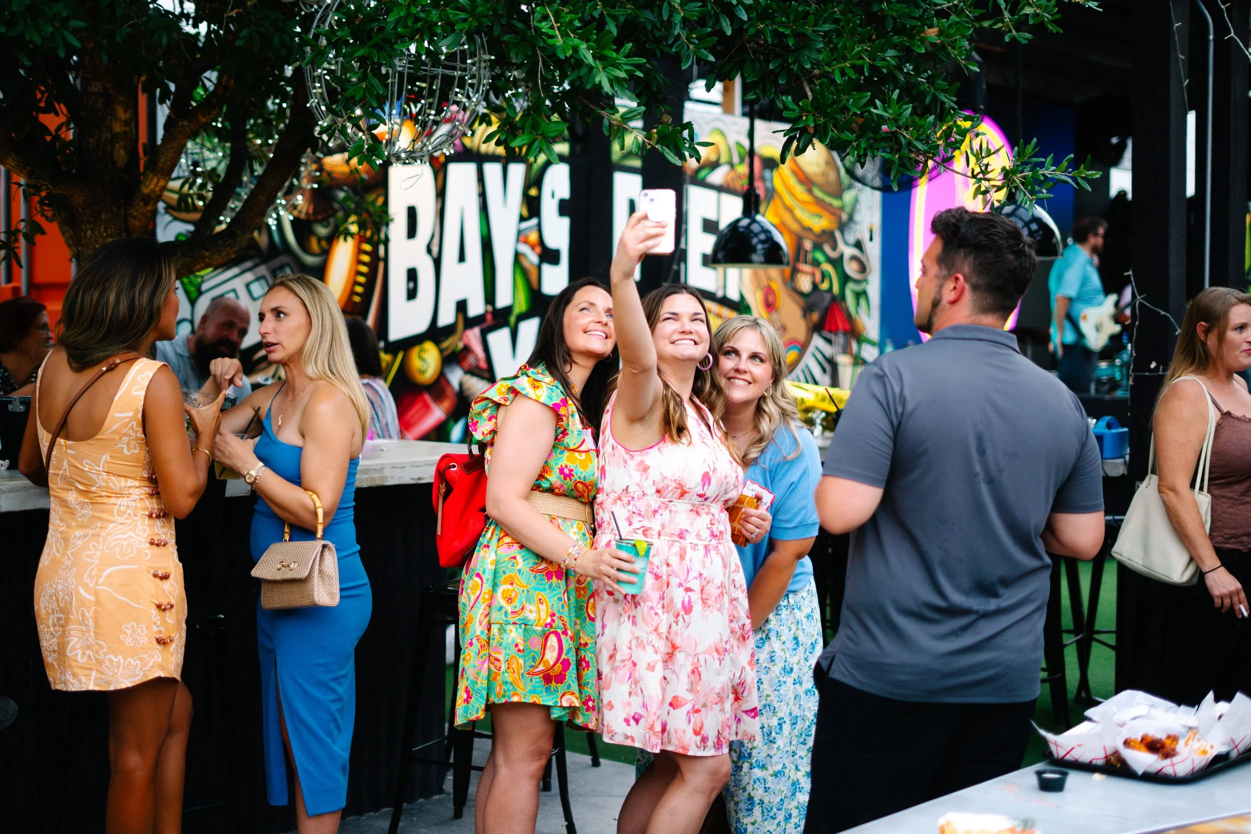 Group of people at a lively outdoor event, some women are smiling and taking selfies, with colorful waves and the word 'BAR' visible in the background.