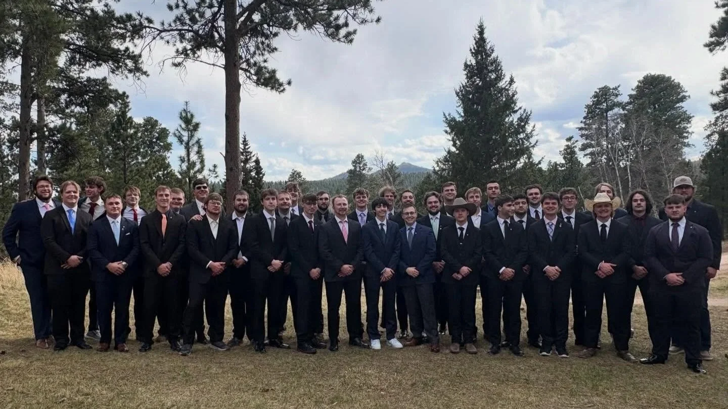 A large group of men dressed in suits posing outdoors in a forested area with trees and mountains in the background.
