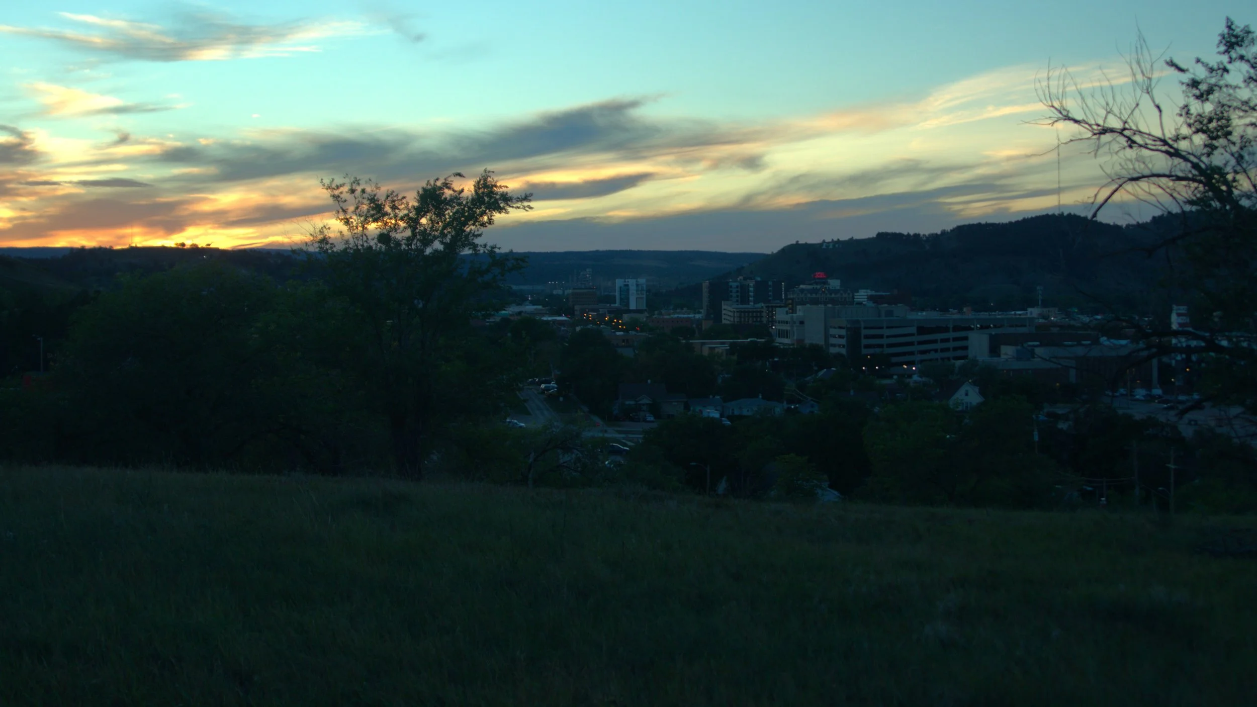 Sunset over a cityscape with buildings and hills, trees in the foreground, and a colorful sky with clouds.