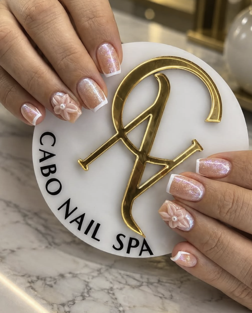 Manos con uñas decoradas con esmalte rosa con destellos y detalles en blanco y rosa con una flor y perla, sosteniendo un letrero redondo con letras doradas que dice 'NY'; en la parte inferior, letrero que dice 'Cabo Nail Spa' en negro.