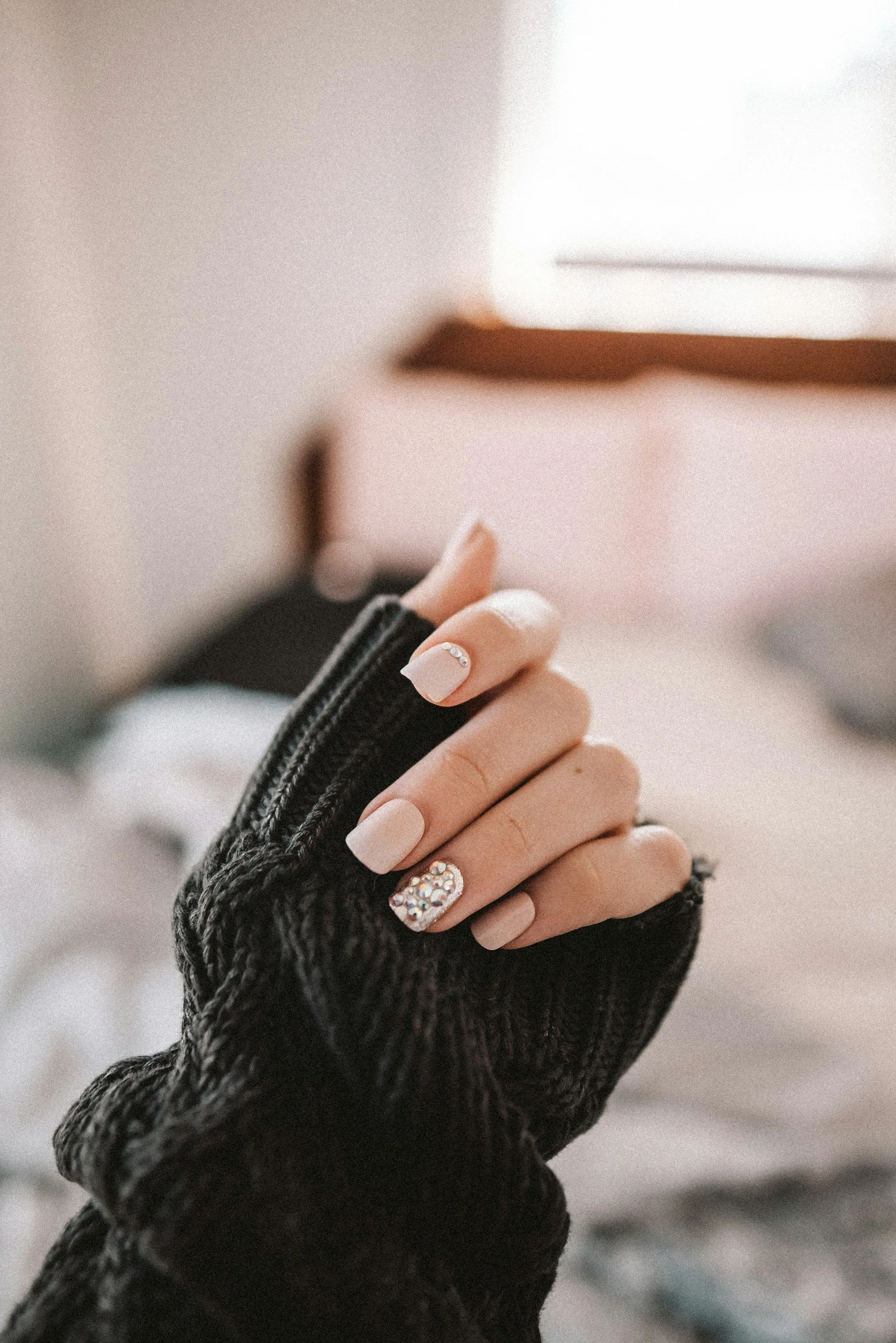 Close-up of a person's hand with manicured nails, some with rhinestone embellishments, wearing a black, textured glove, and holding what appears to be a small mirror or compacted object. The background is softly blurred.