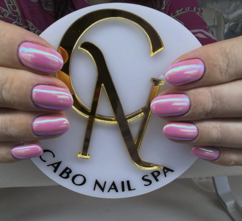 Manicura con uñas largas pintadas de color rosa con detalles en azul y verde en una mano, sosteniendo un letrero redondo con letras doradas y negras que dice "Cabo Nail Spa".