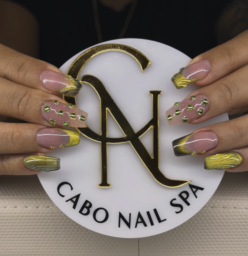 Manicure con uñas decoradas con esmalte en tonos oro, gris y verde, con brillo y piedras pequeñas, sosteniendo un logo de cabo nail spa.