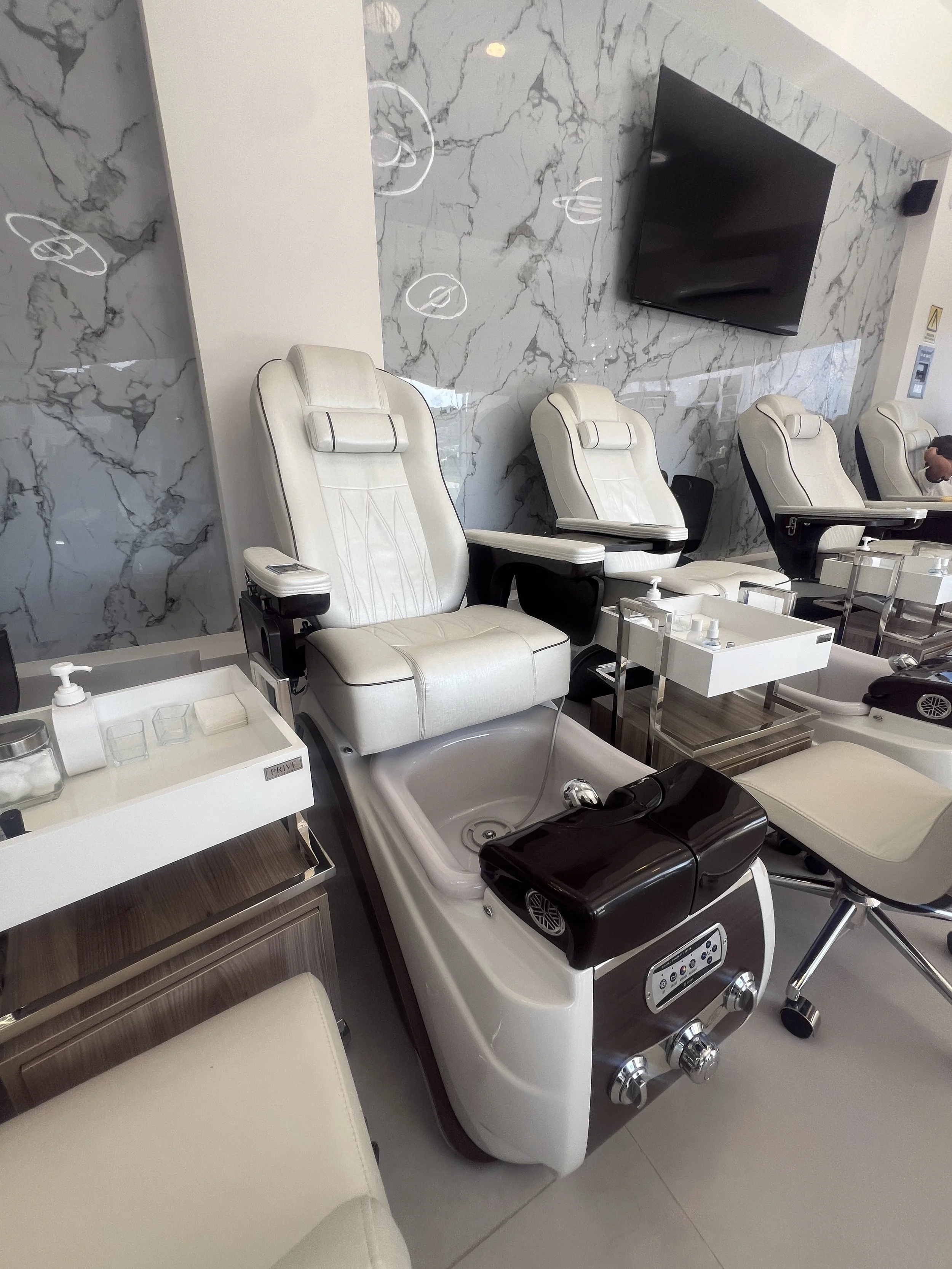 Sala de espera en un salón de belleza o spa con sillas blancas de masaje, mesas con productos y una pared de mármol gris con negro. En la pared hay un televisor y a la derecha hay un altavoz y una persona sentada.