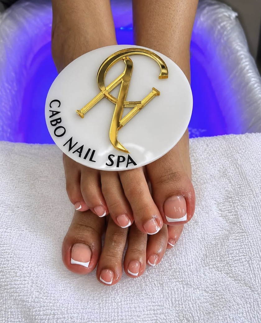 Manos con uñas arregladas, descansando sobre una toalla blanca en un spa. Sobre las manos hay un parche con el logo de Cabo Nail Spa, que es de color blanco con letras negras y detalles dorados.