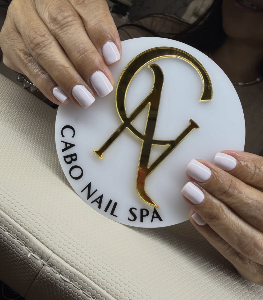 Manos con uñas pintadas de blanco sosteniendo un cartel redondo que dice 'Cabo Nail Spa' y tiene un logo dorado con las letras 'C' y 'V' entrelazadas.