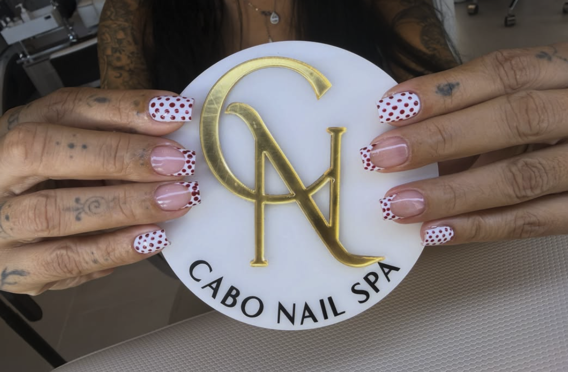 Mano con uñas decoradas con esmalte rojo con puntos blancos, sosteniendo un logotipo dorado y blanco del spa de uñas Cabo Nail Spa.