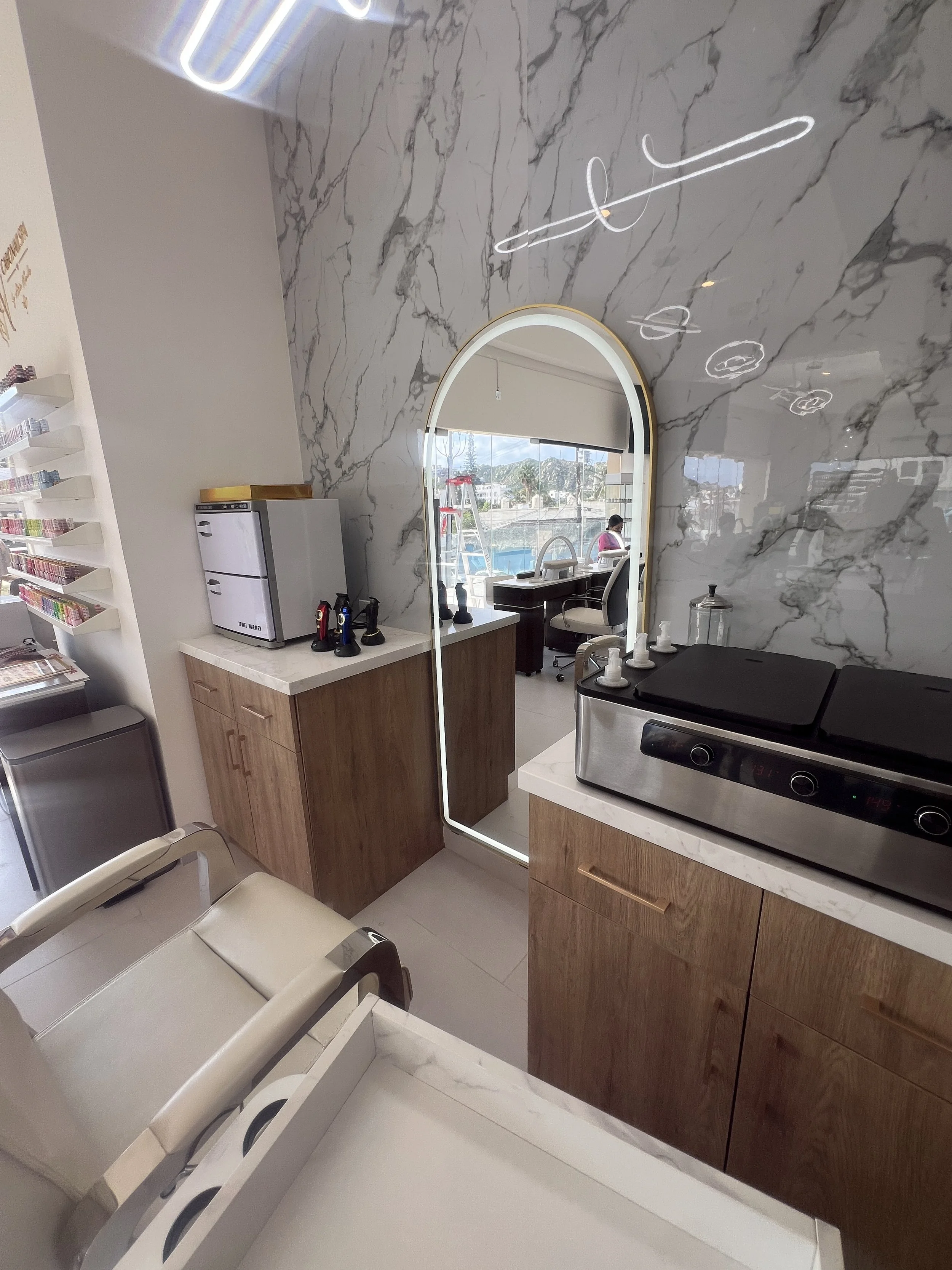 Interiores de una clínica o consultorio con pared de mármol, espejo con luz, mesa con equipo de café y taza, área de espera con sillas blancas, y vista de un patio con piscina a través de la ventana.