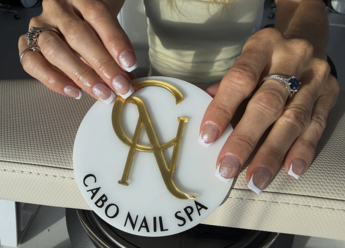 Manos con uñas pintadas de blanco y brillo, sosteniendo un letrero de madera con letras doradas, que dice 'Cabo Nail Spa'.