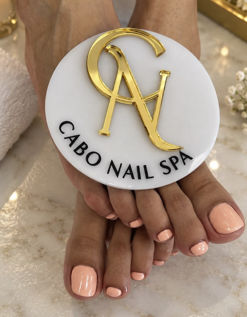 Pendientes de uñas de color melocotón en manos con fondo de spa y logo de Cavo Nail Spa en una placa de mármol blanco con letras doradas y negras.