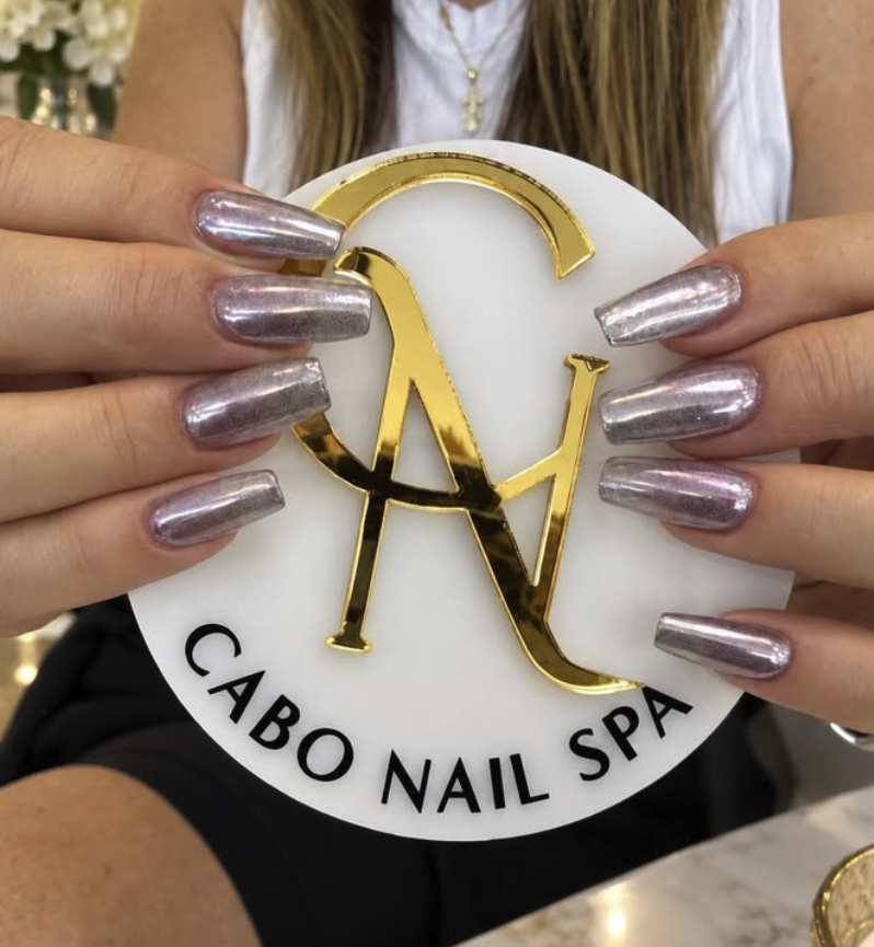Mano con uñas largas y brillantes sosteniendo un logo dorado en forma de letras "C" y "N" sobre un letrero que dice "Cabo Nail Spa."