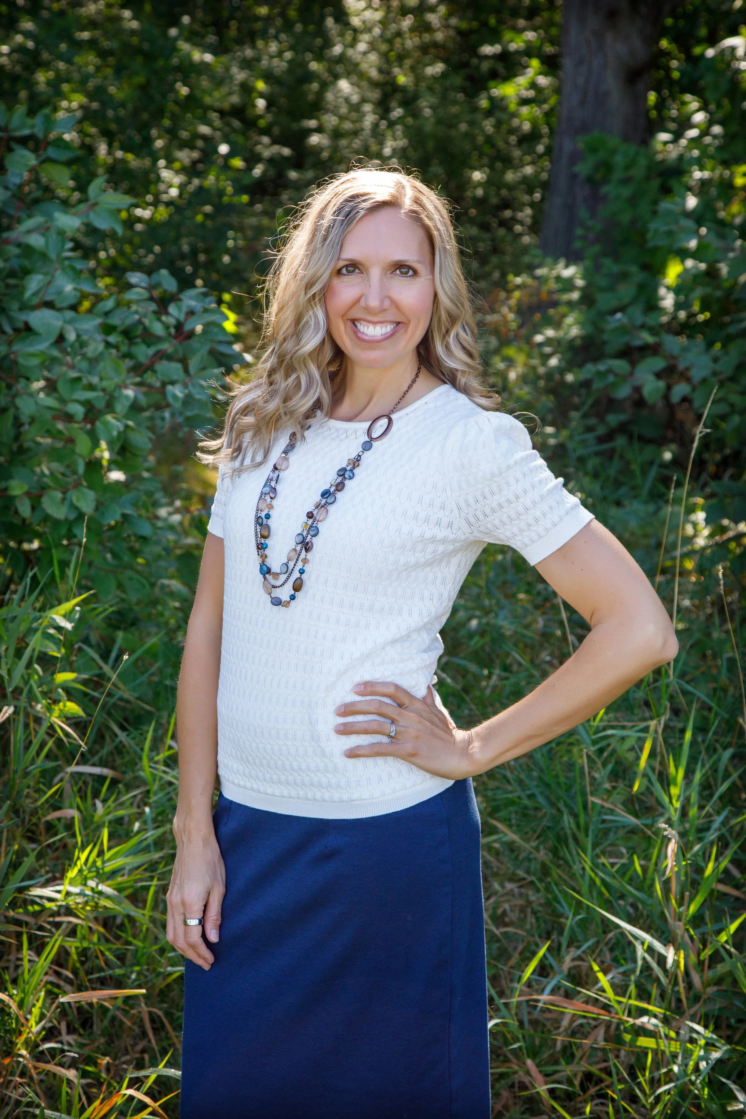 A woman with blonde curly hair, smiling, standing outdoors among greenery. She is wearing a white short-sleeved shirt, a multi-strand necklace, and a dark blue skirt.