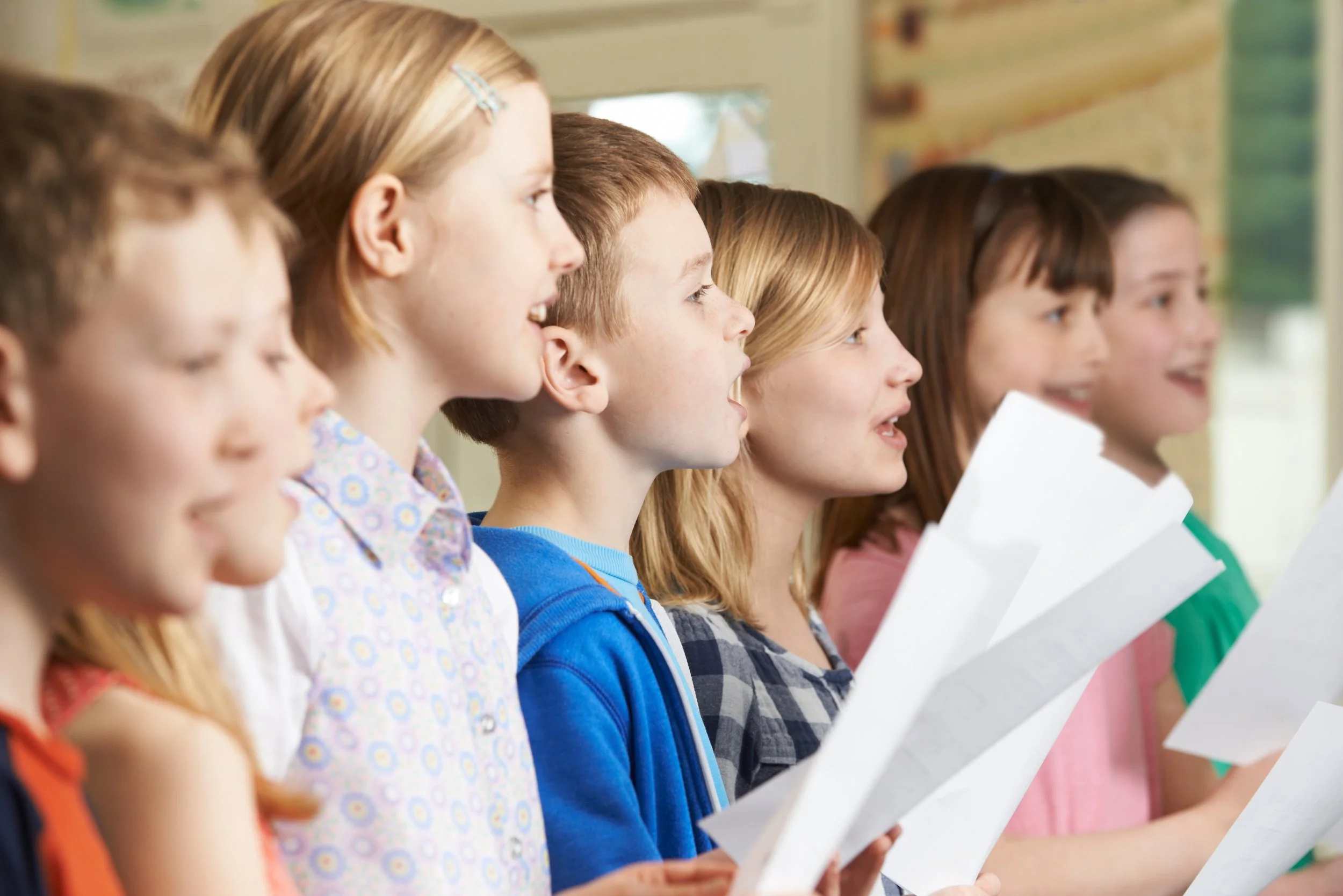 A row of children standing side by side, holding music, participating in a singing or presentation activity inside a classroom with large windows and colorful walls.