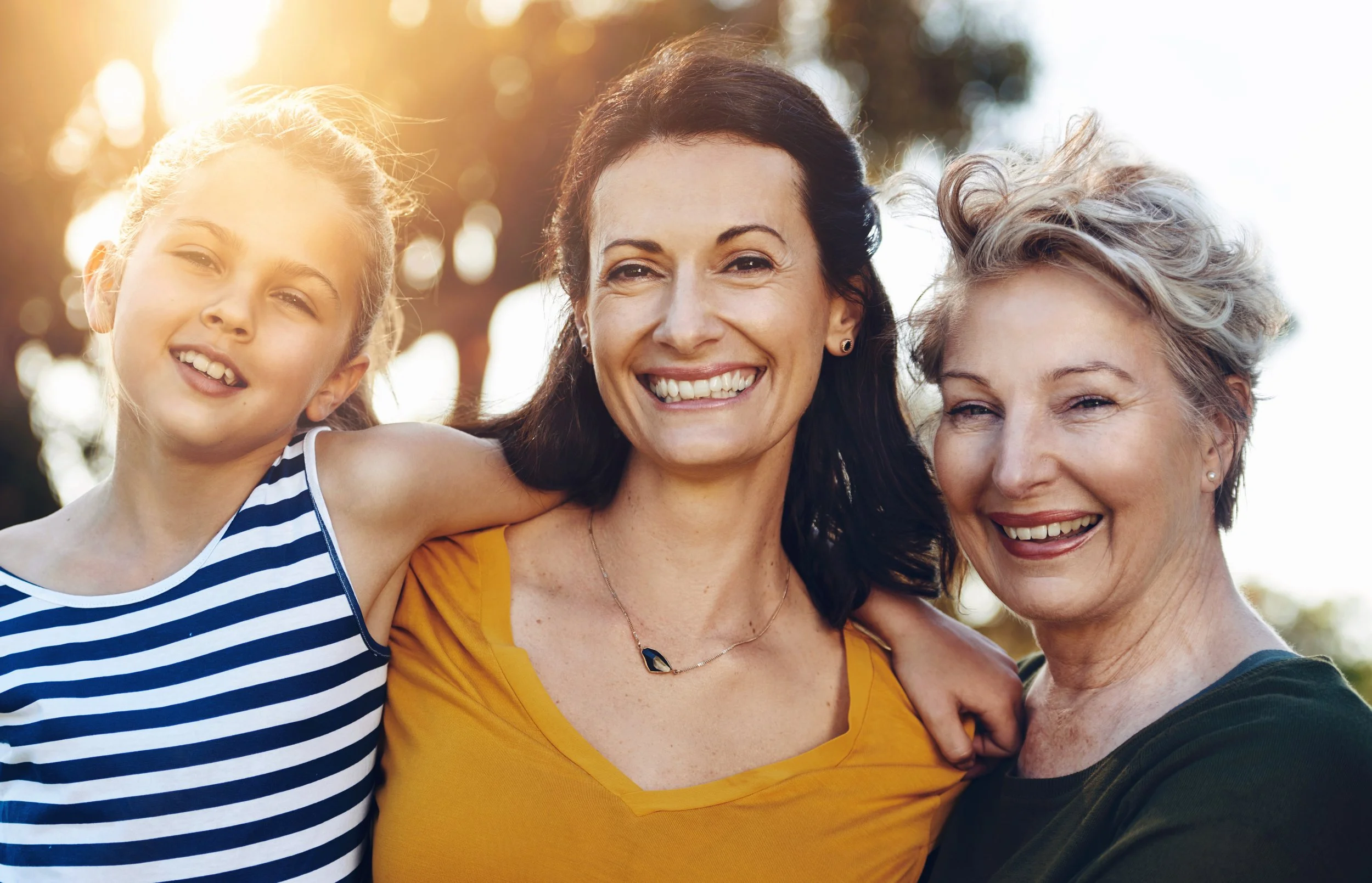 Three women and a girl smiling outdoors during sunset, with trees in the background.