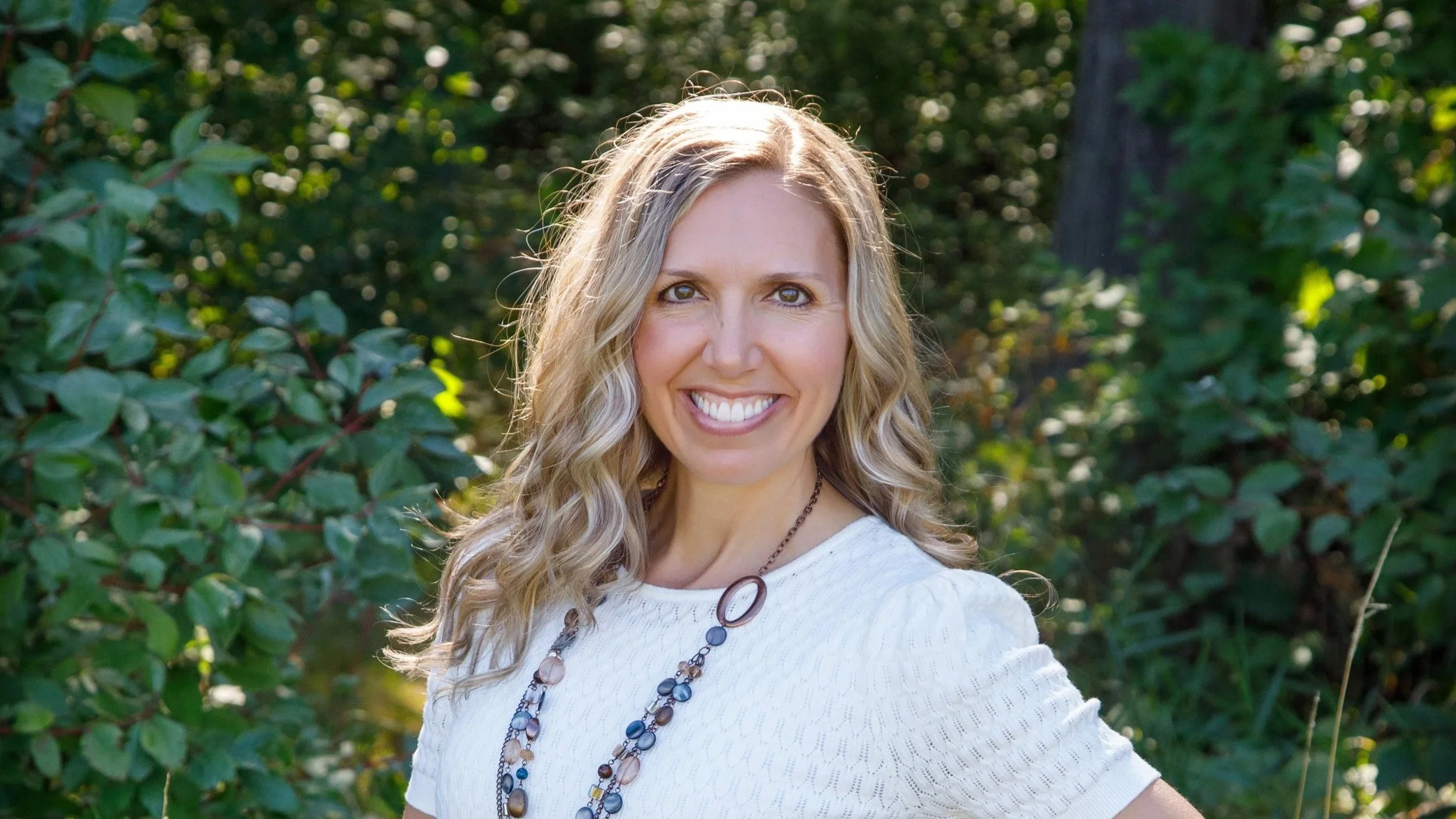 Portrait of a smiling woman with wavy blonde hair, wearing a white top and a multi-colored beaded necklace, outdoors with green foliage in the background.