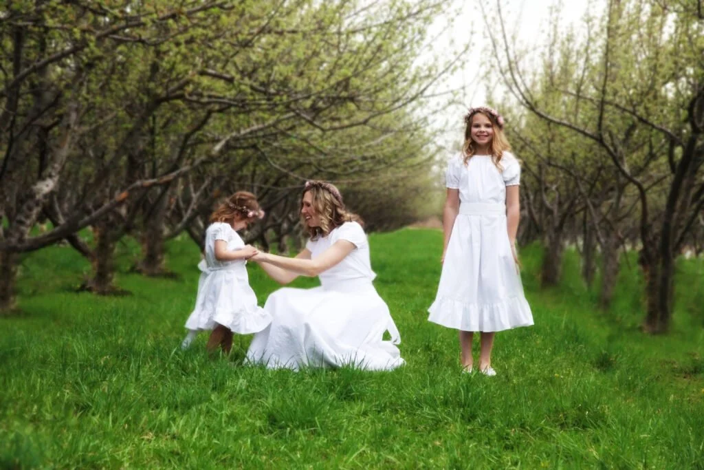 A woman and two young girls in white dresses in an orchard