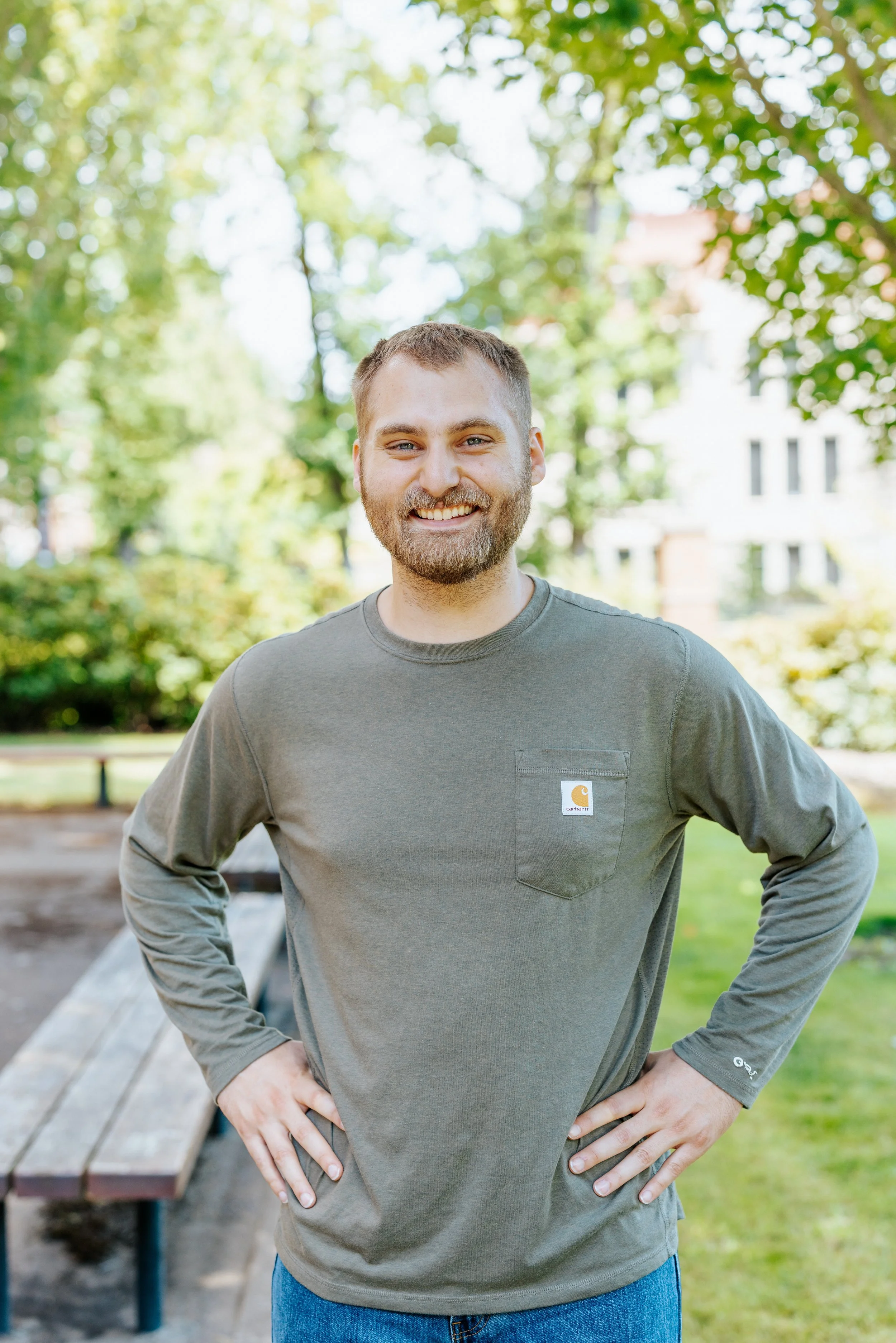 A smiling man with a beard and short hair standing outdoors at a park, wearing a gray Carhartt long-sleeve shirt and blue jeans, with his hands on his hips. Background includes trees and a building.