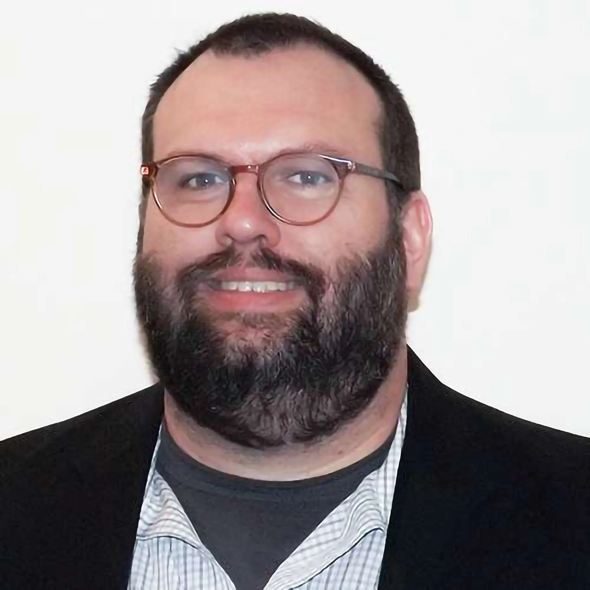 A man with glasses, a beard, and short hair wearing a black jacket and white collared shirt, smiling against a white background.