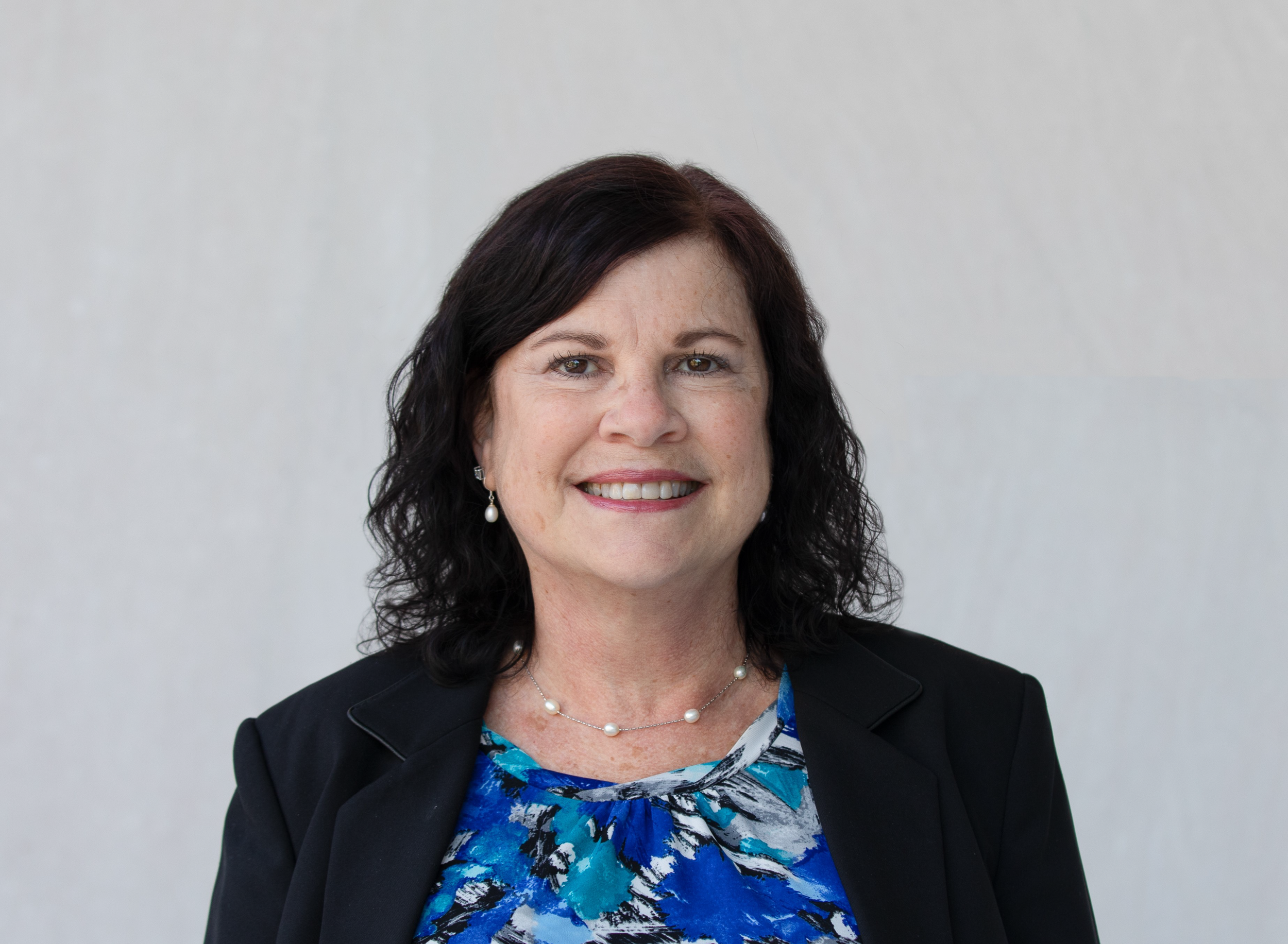 Close-up of a smiling woman with dark brown hair, wearing a black blazer, pearl earrings, a pearl necklace, and a blue patterned blouse against a plain light gray background.
