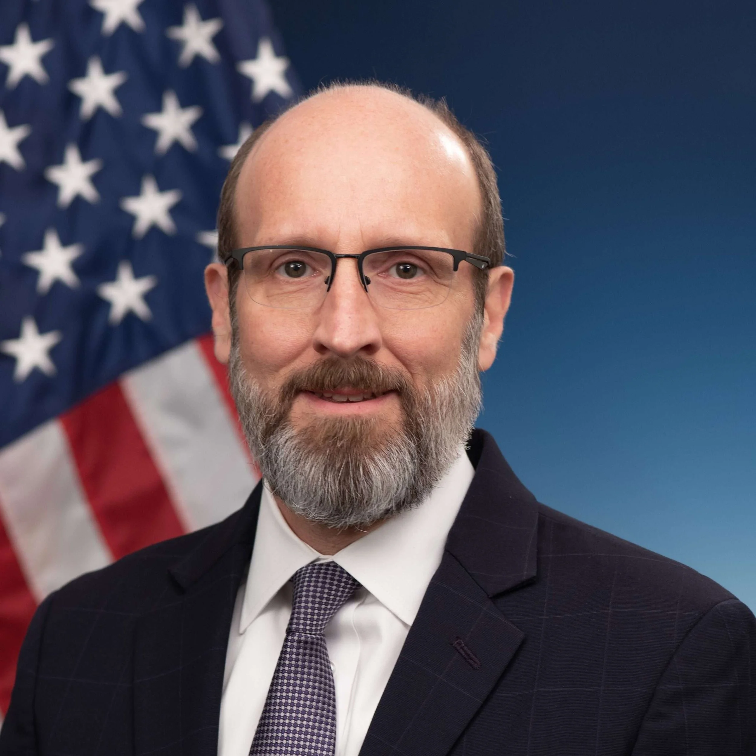 Close-up of a middle-aged man with a beard and glasses, wearing a suit and tie, standing in front of an American flag background.