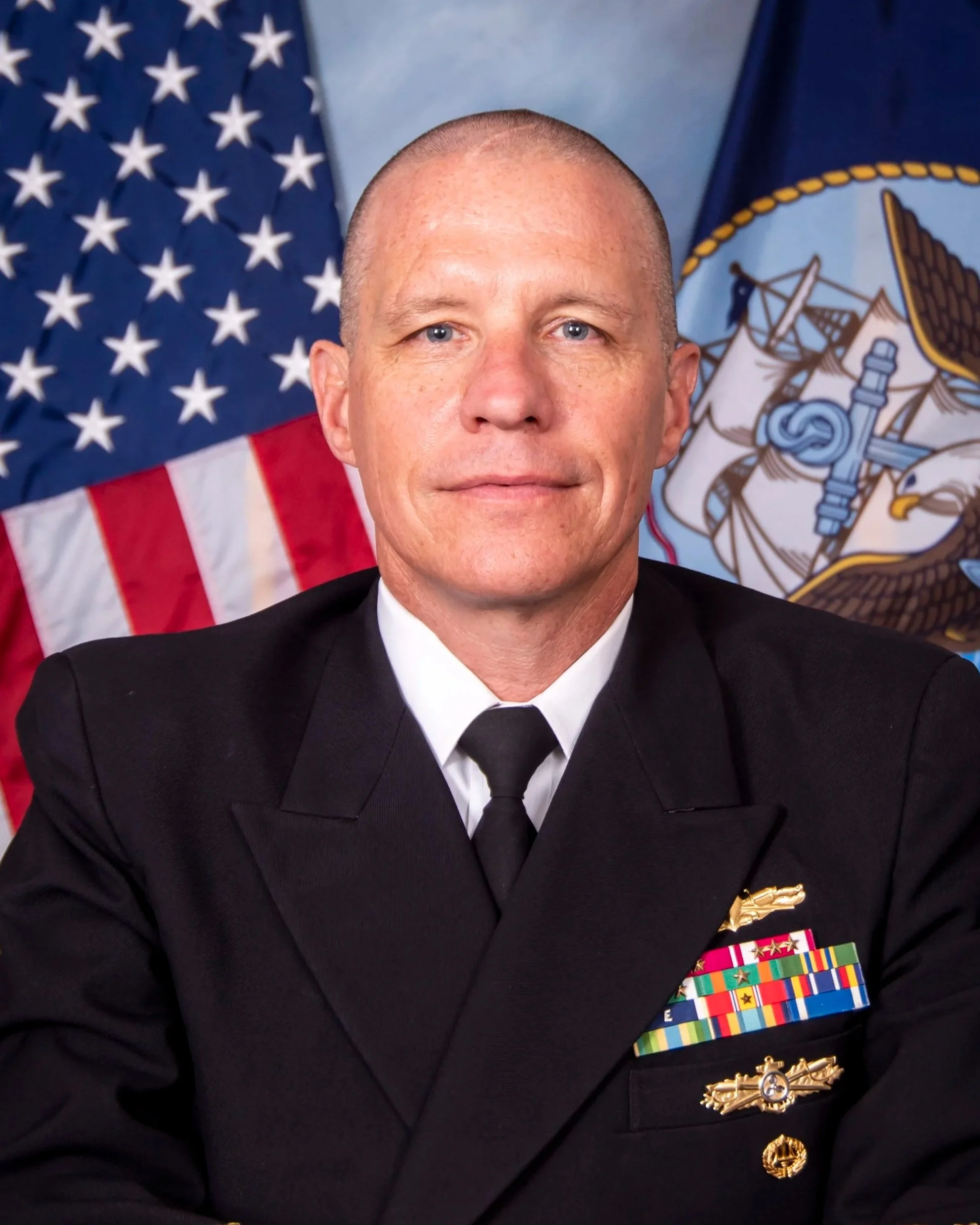 A man in a military uniform with numerous ribbons and medals, sitting at a table with his hands clasped, flanked by American and military flags in the background. US Navy Admiral. 