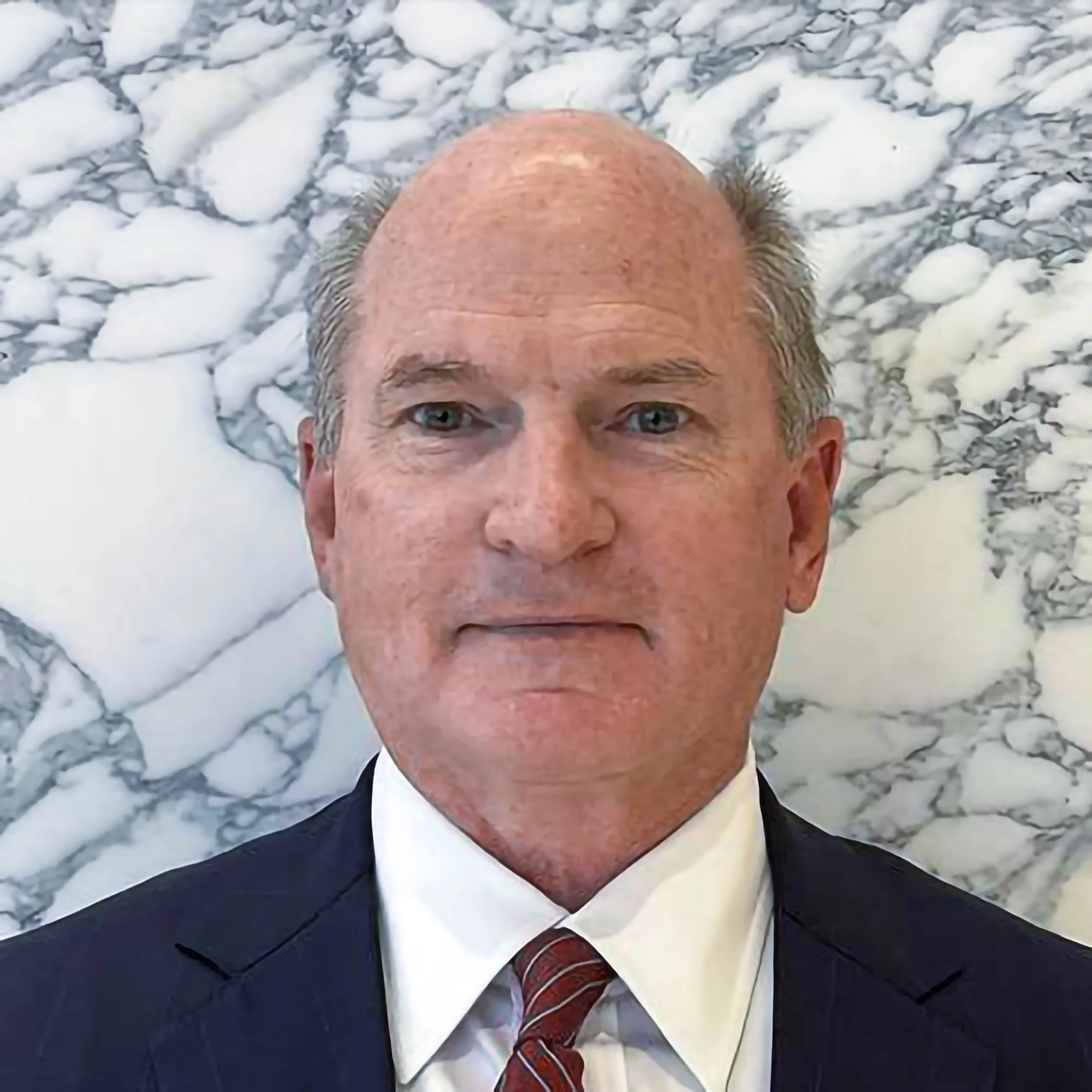 Close-up of a middle-aged man with light skin, blue eyes, and a bald head, smiling slightly and wearing a dark suit with a red patterned tie, lying on a marble surface.