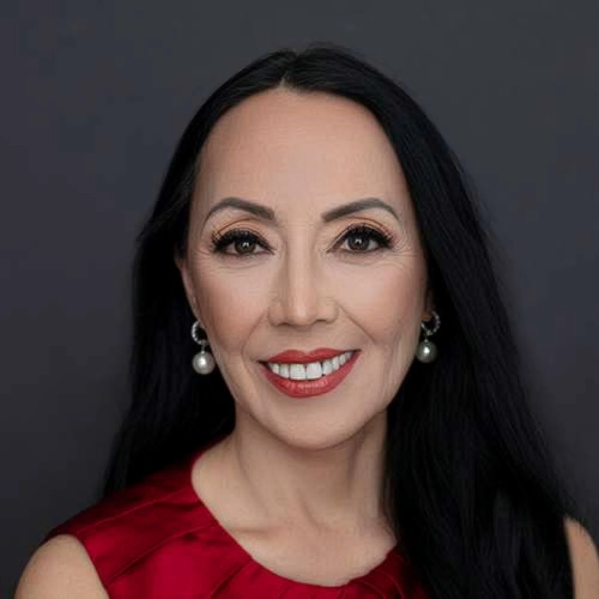 Close-up of a woman with long black hair, wearing pearl earrings and a red dress, smiling.