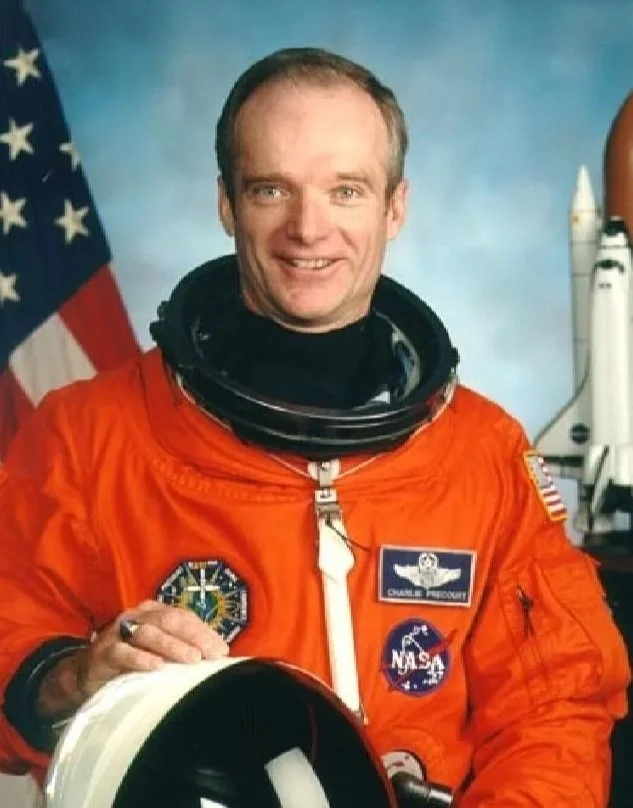 A man dressed in a bright orange NASA astronaut suit, standing in front of an American flag, smiling at the camera. Charlie Precourt. 