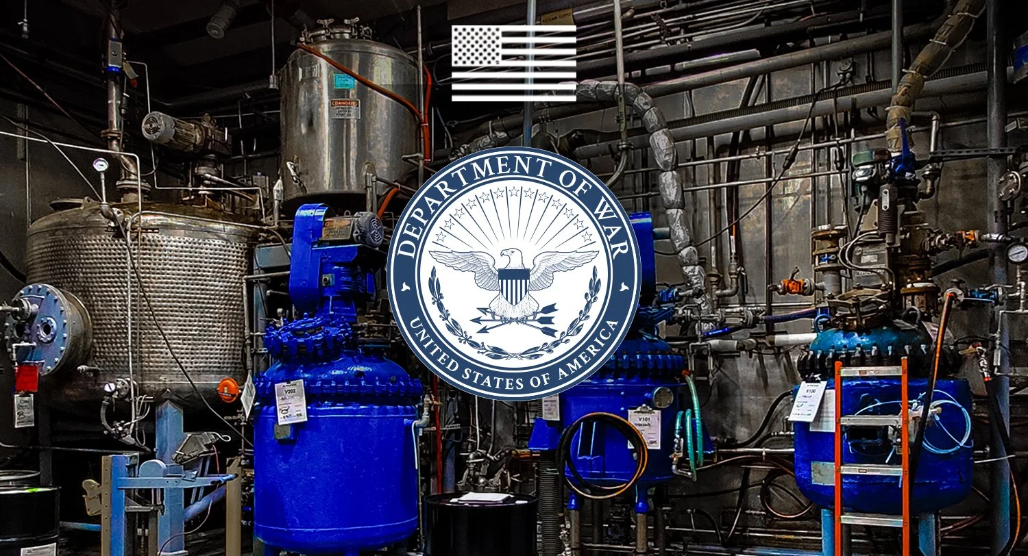 Industrial equipment and machinery behind the emblem of the Department of War, United States of America, with the American flag above.