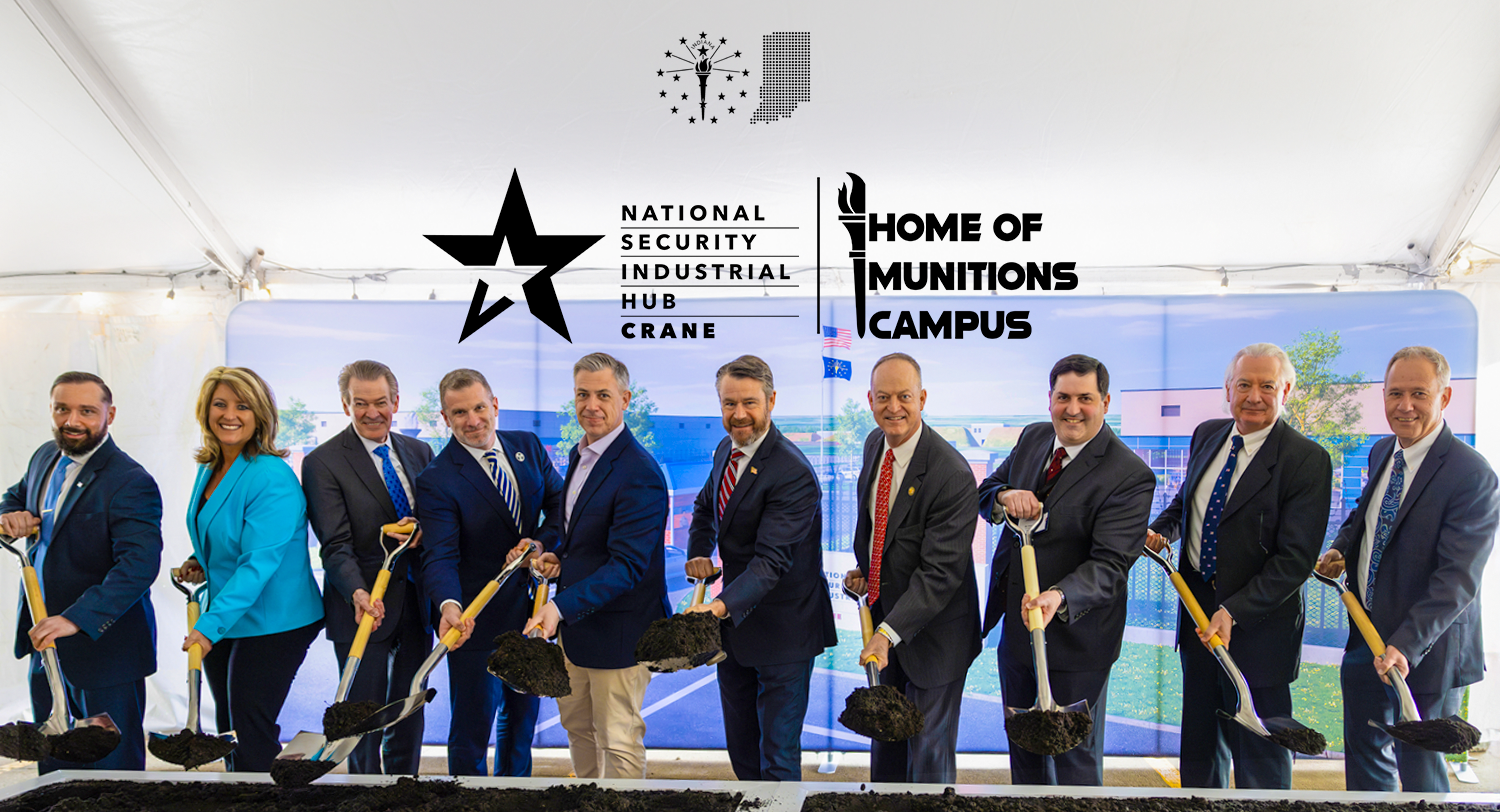 A group of ten people in business attire participating in a groundbreaking ceremony, each holding a small shovel with dirt, with logos and banners of the National Security Industrial Hub Crane and the Home of Munitions Campus in the background.