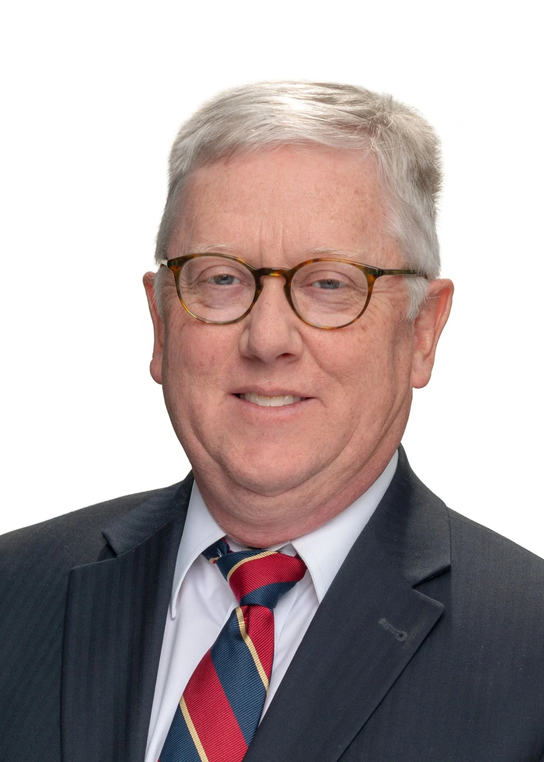 Close-up of a smiling older man with gray hair and glasses, wearing a dark suit, white shirt, and a red, blue, and yellow striped tie, against a white background.