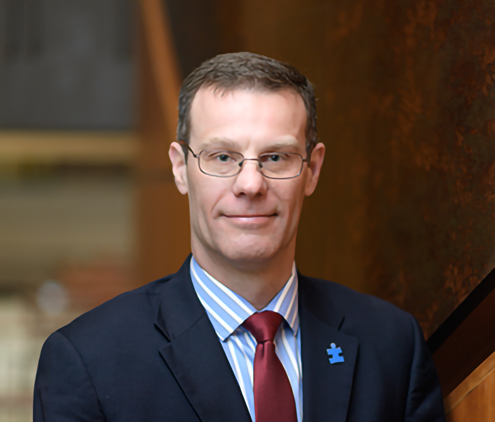 A man with glasses, wearing a dark suit, a striped shirt, and a red tie, standing indoors with a wooden background.