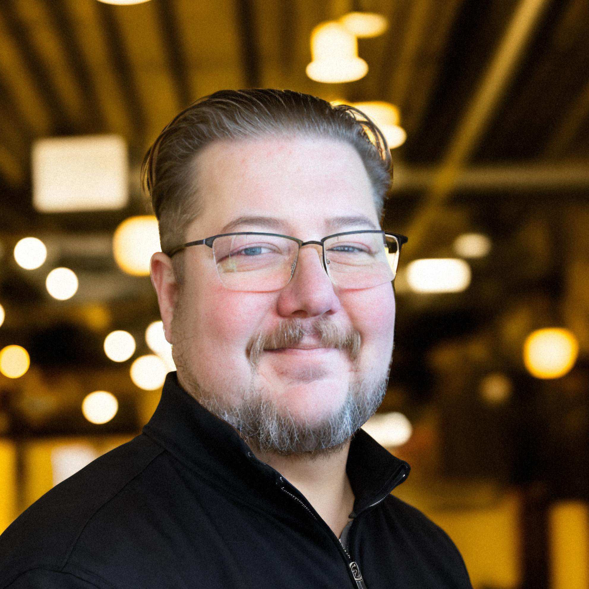 Close-up of a man with glasses and facial hair, wearing a black jacket, in front of a background with warm yellow and orange lights.