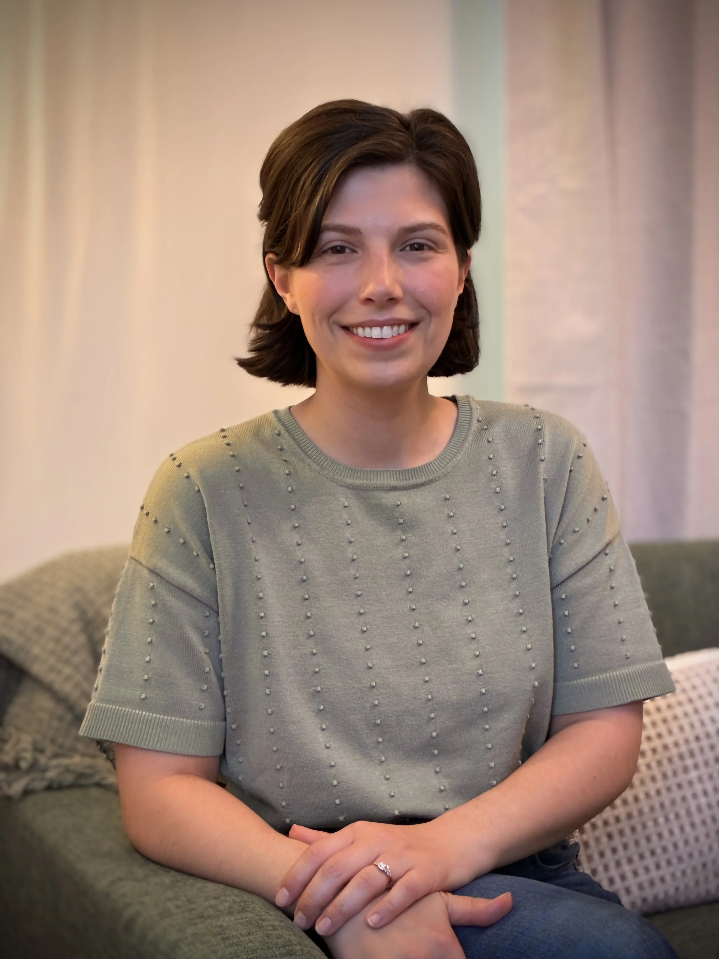 A woman with short brown hair smiling, sitting on a green couch with her hands resting on her lap.
