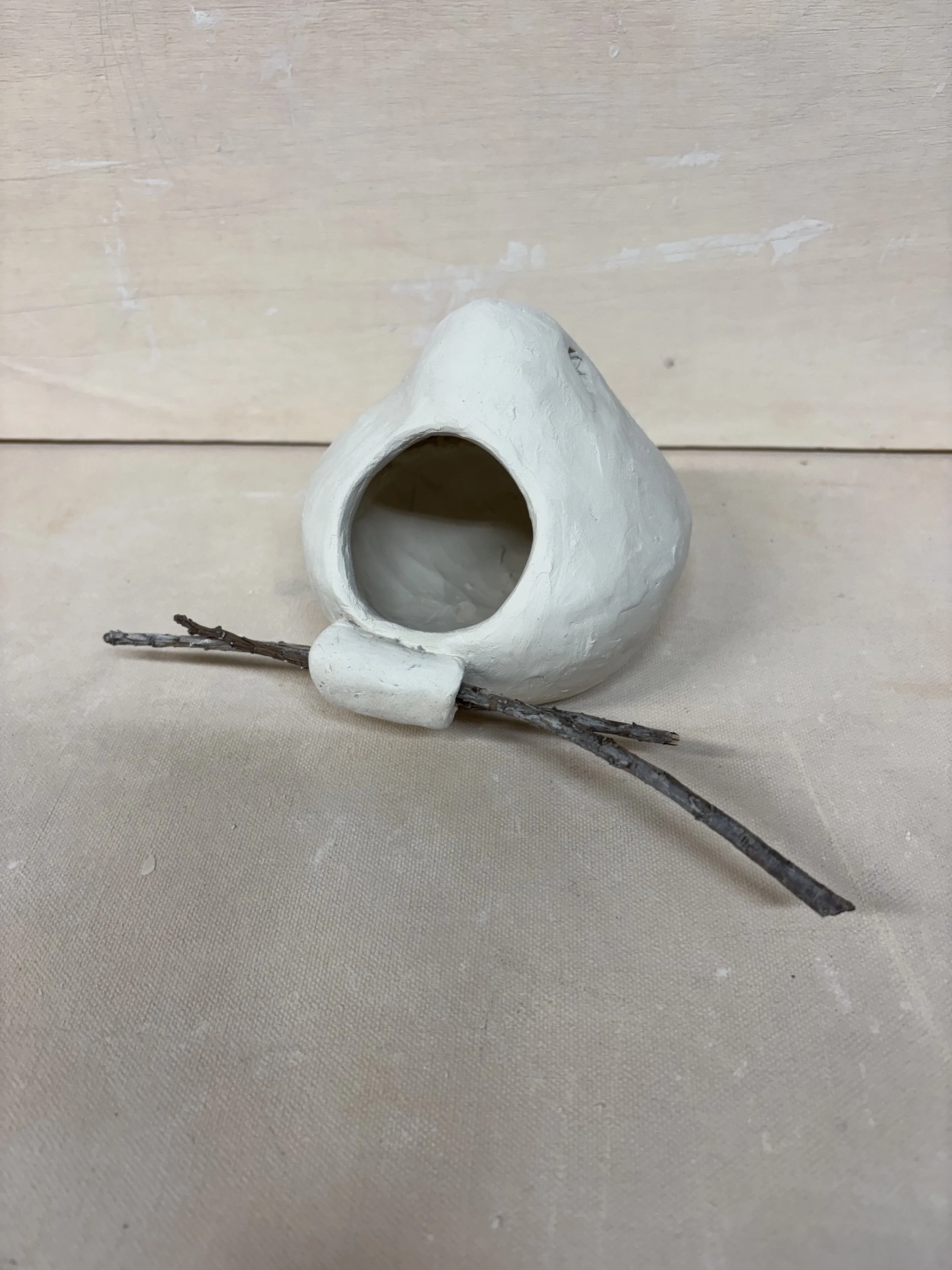 A small, white, ceramic birdhouse with a round entrance hole, placed on a beige fabric surface, with a small twig resting in front of it.