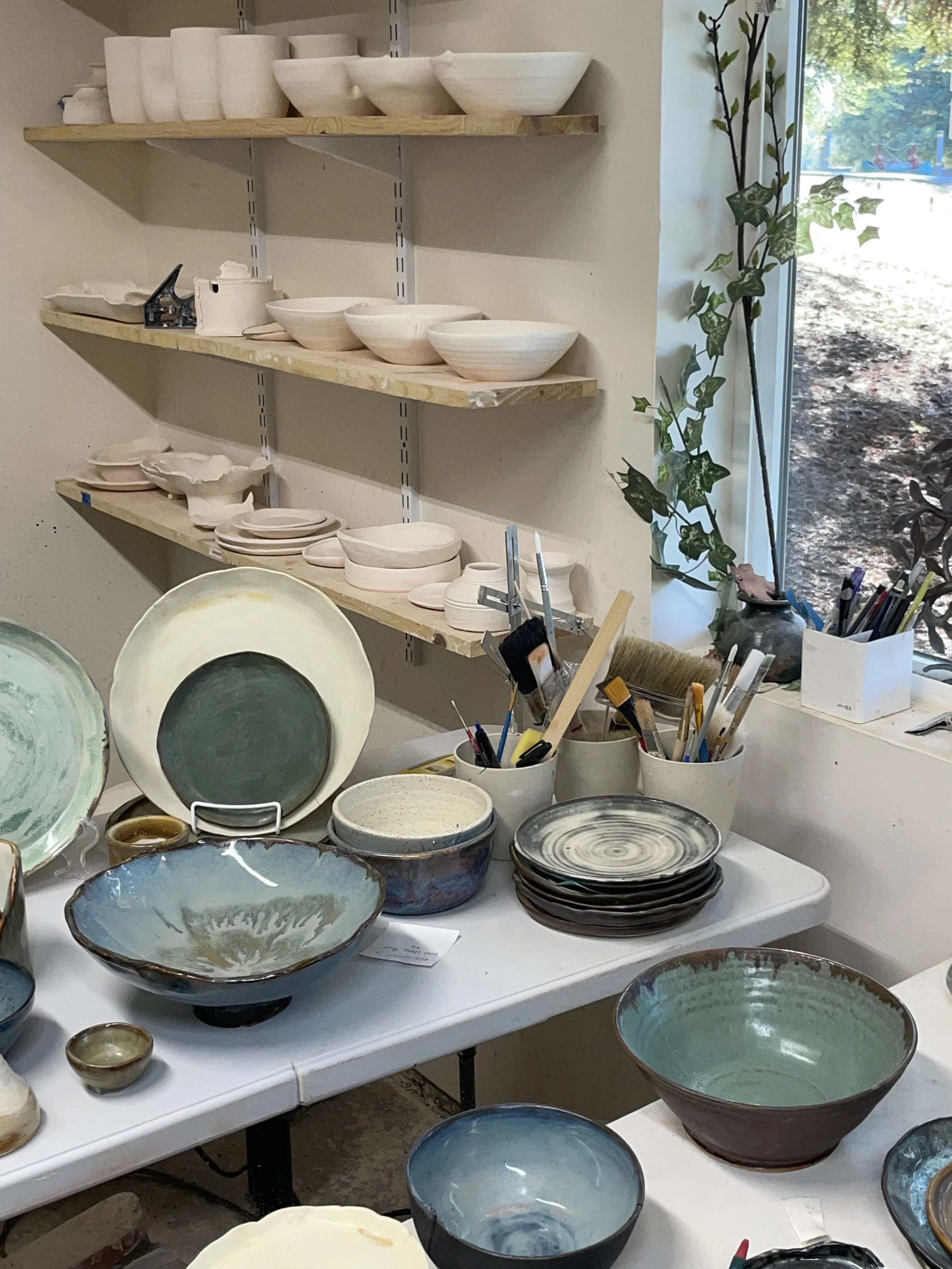 A work table displaying various ceramic bowls and plates in different sizes and colors, with shelves holding more ceramics, and a window with a view of trees outside.