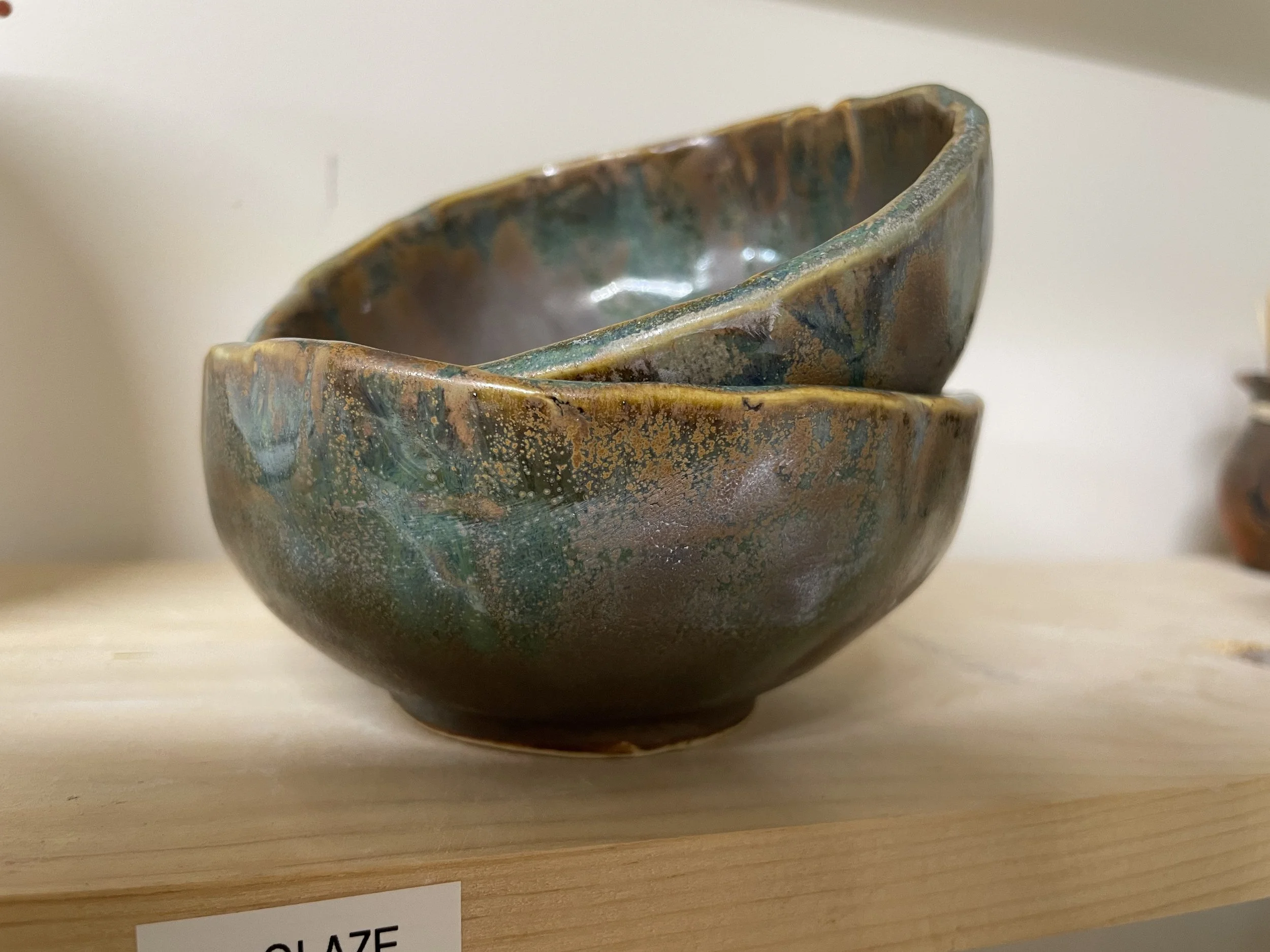Close-up of a textured ceramic bowl with a mottled green, brown, and blue glaze, sitting on a light wooden surface.
