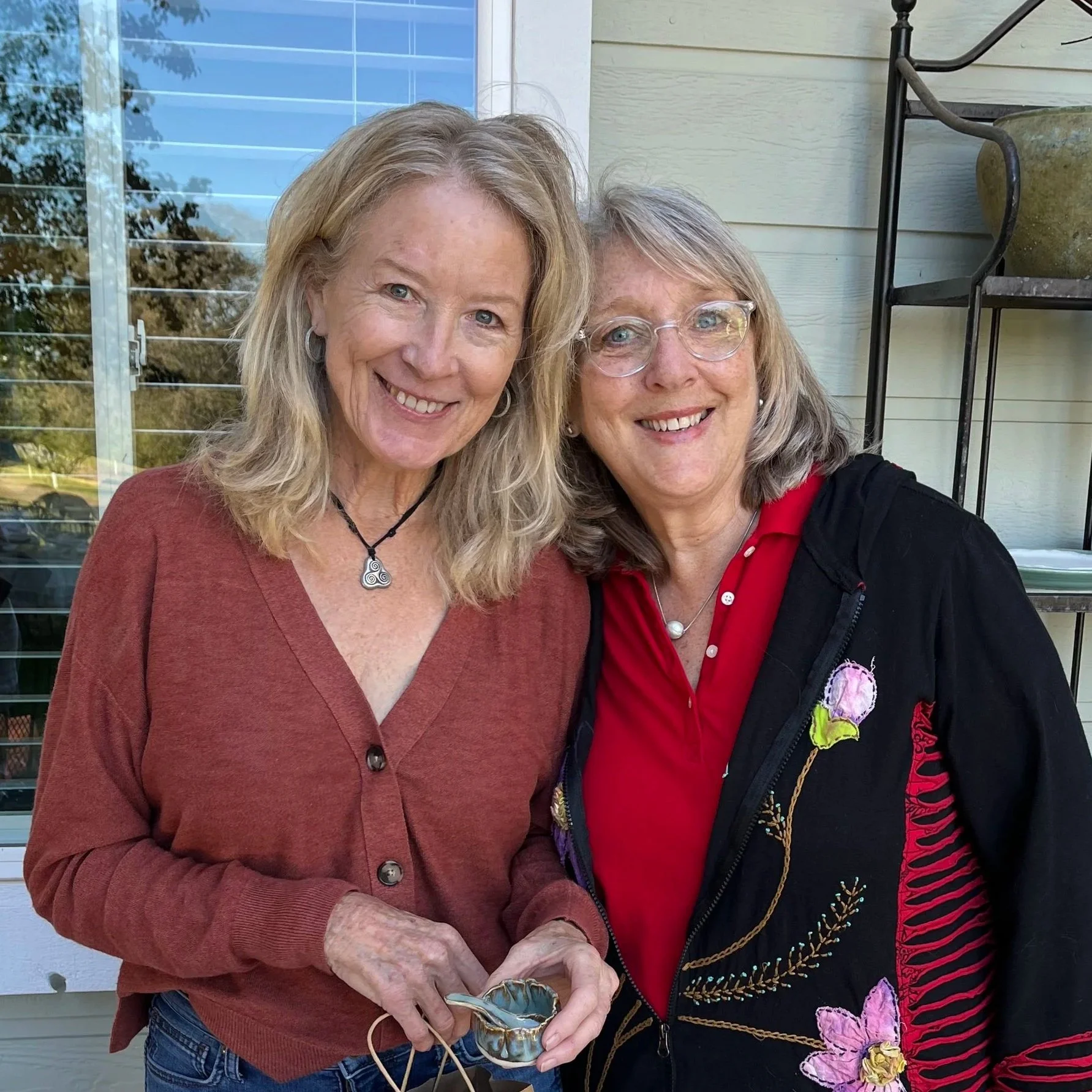 Two women smiling outdoors, one with blonde hair wearing a red blouse with a black jacket, the other with gray hair and glasses wearing a red shirt and black jacket with floral embroidery.
