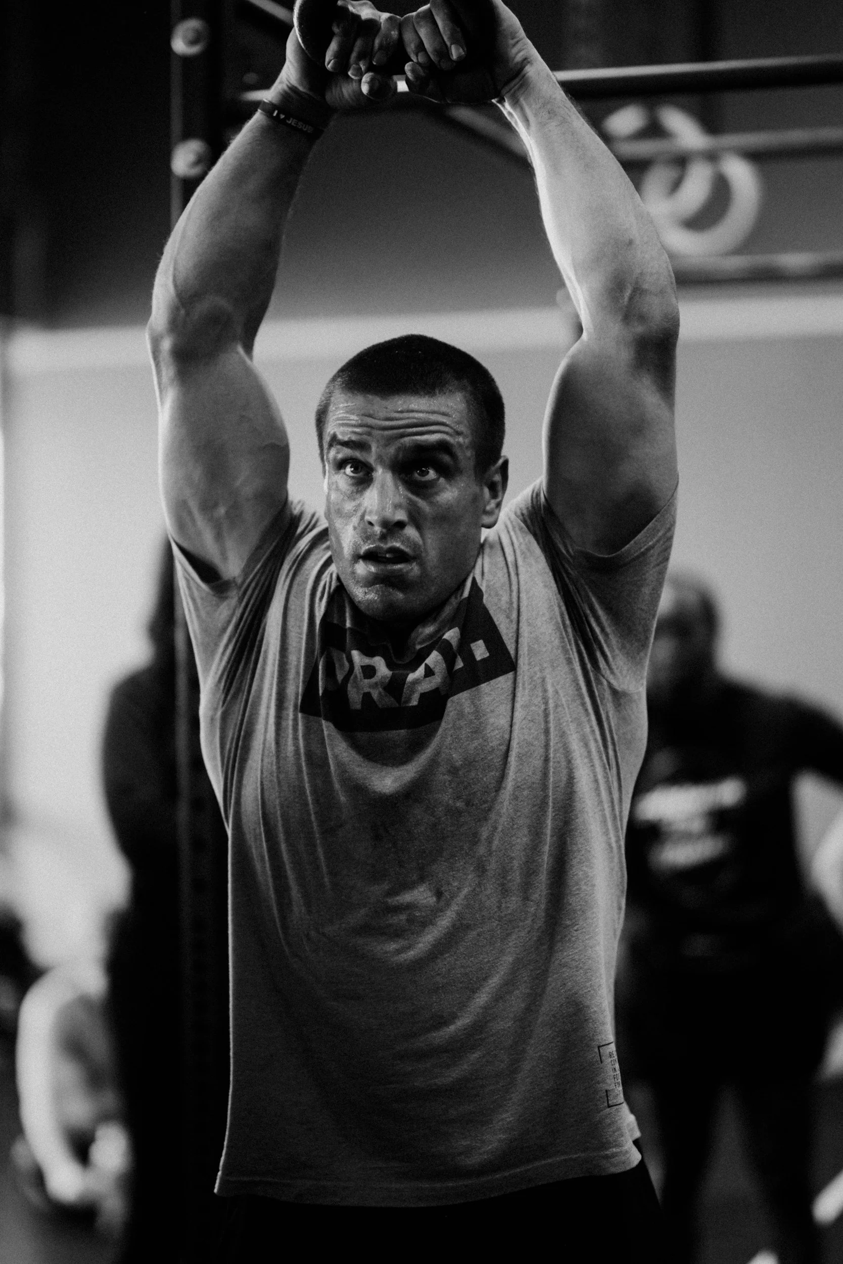 A man with short hair and a serious expression is standing with his arms raised above his head and hands clasped together. He is wearing a T-shirt and a wristband, and appears to be in a gym or fitness center.