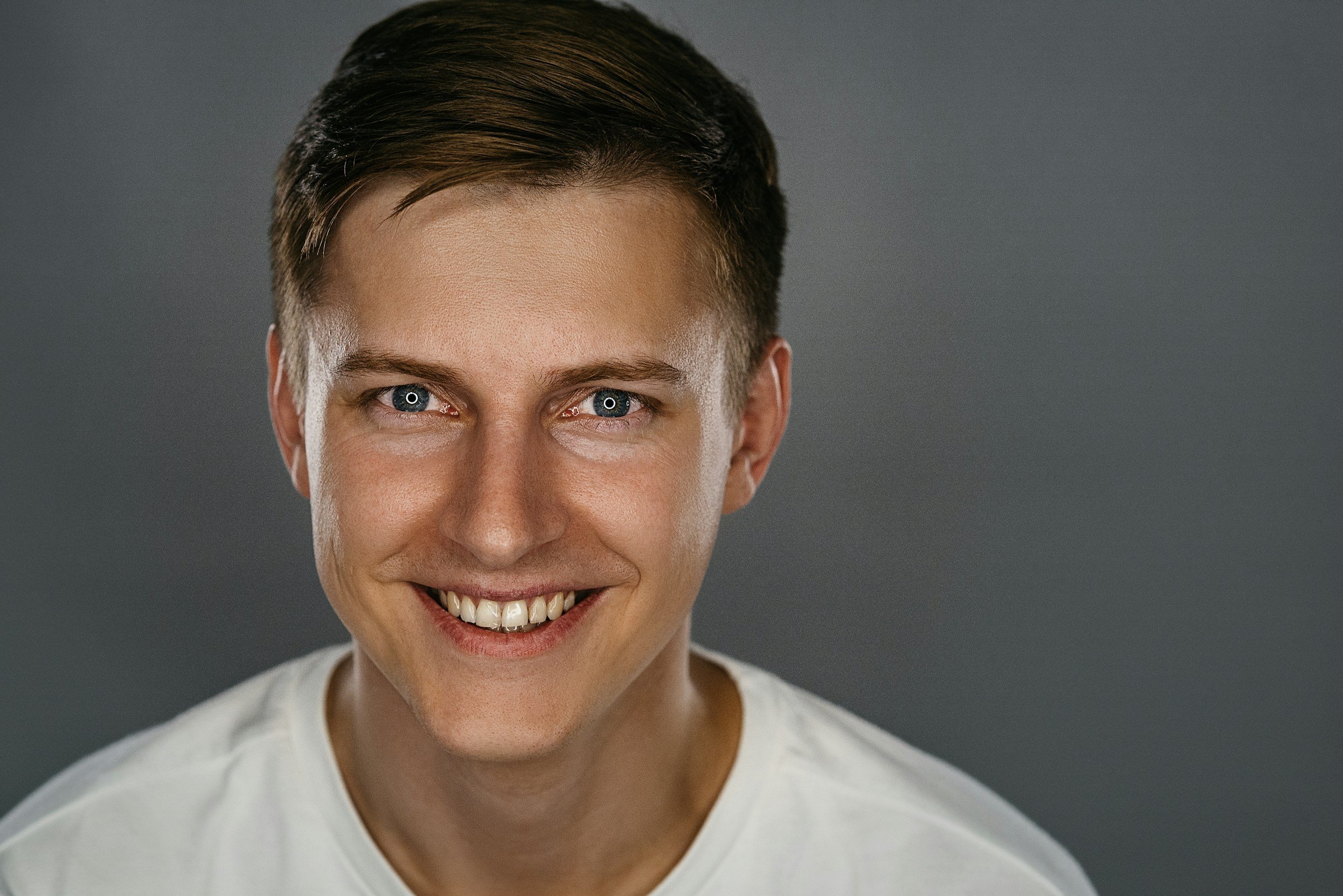Close-up of a smiling person with short brown hair and blue eyes, wearing a white shirt, against a gray background.