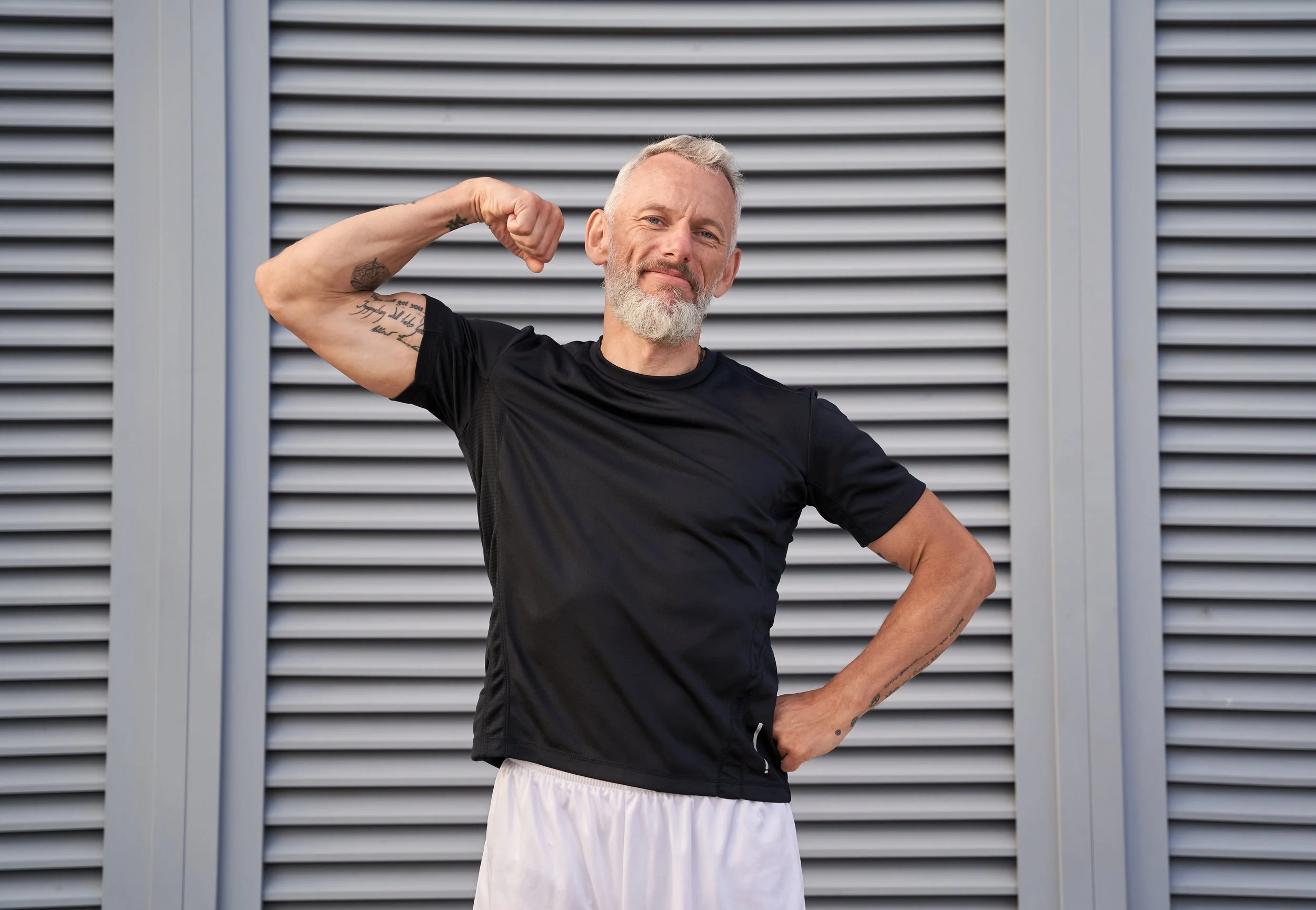 A man with a gray beard and short hair flexes his right arm showing his bicep, standing in front of a modern metal fence wearing a black T-shirt and white shorts.