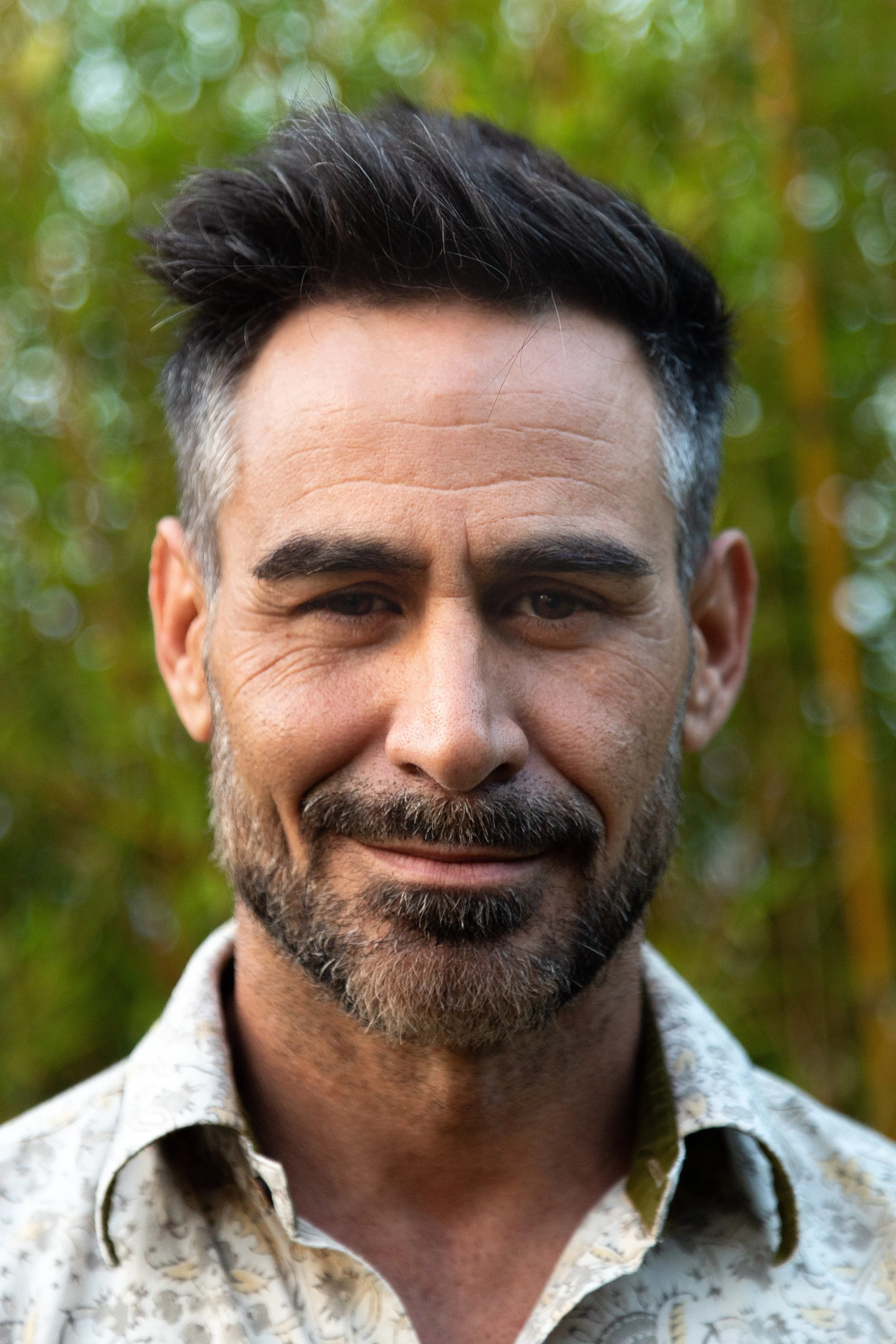 A close-up of a smiling middle-aged man with dark hair, a beard, and a mustache, wearing a patterned shirt, standing outdoors with blurred green foliage in the background.