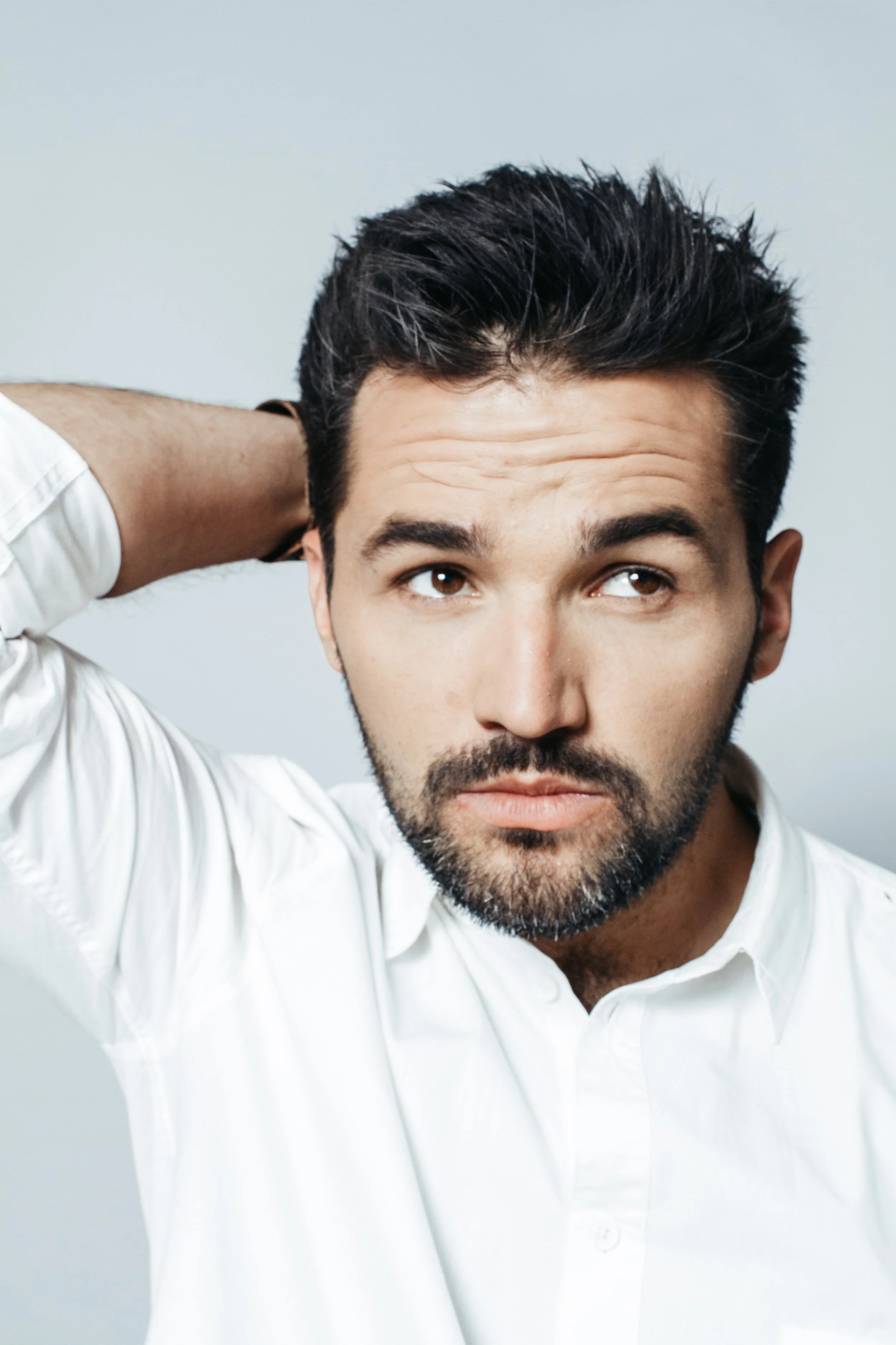 A man with dark hair and a beard wearing a white shirt, looking off to the side and resting his hand on the back of his head.